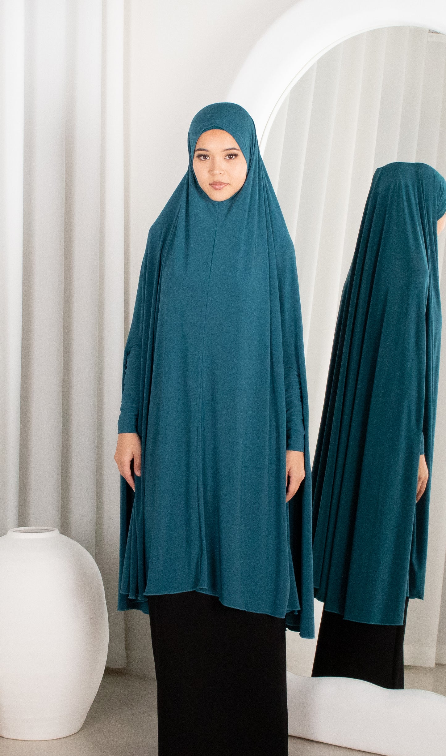 Woman wearing a teal hijab and dress standing in front of a mirror.