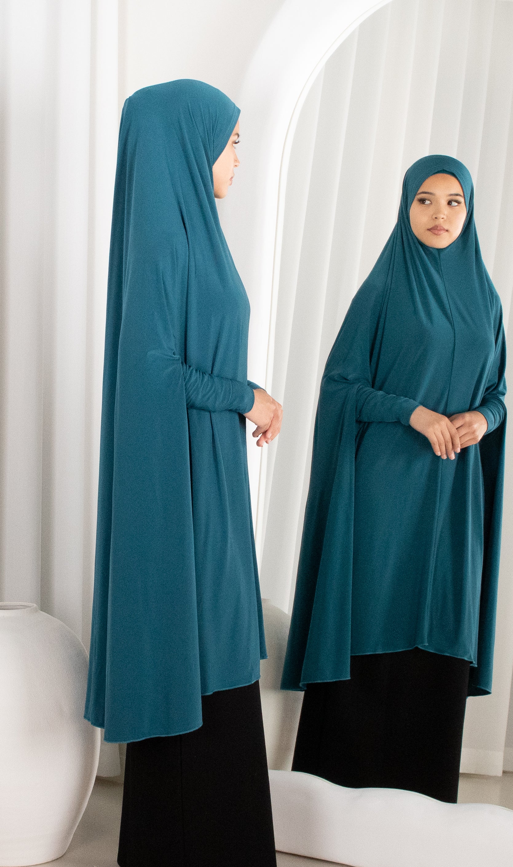 Woman wearing a teal hijab and black outfit standing in front of a white curtain.