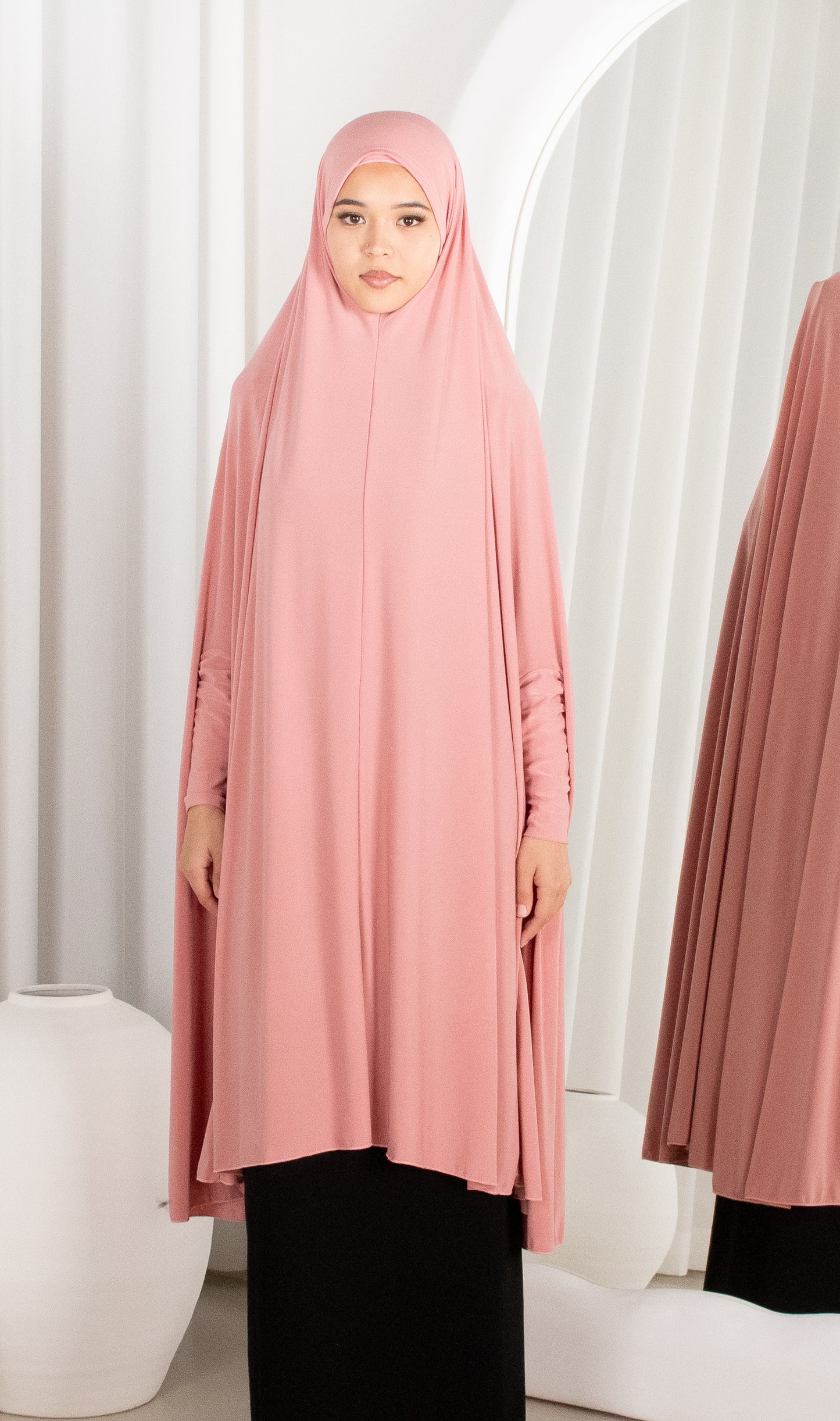 Woman wearing a pink hijab and dress standing against a white curtain.