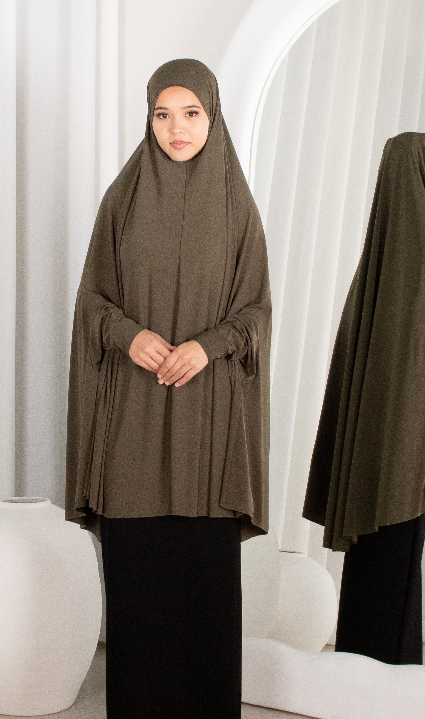 Woman wearing a brown hijab and black outfit in a white room