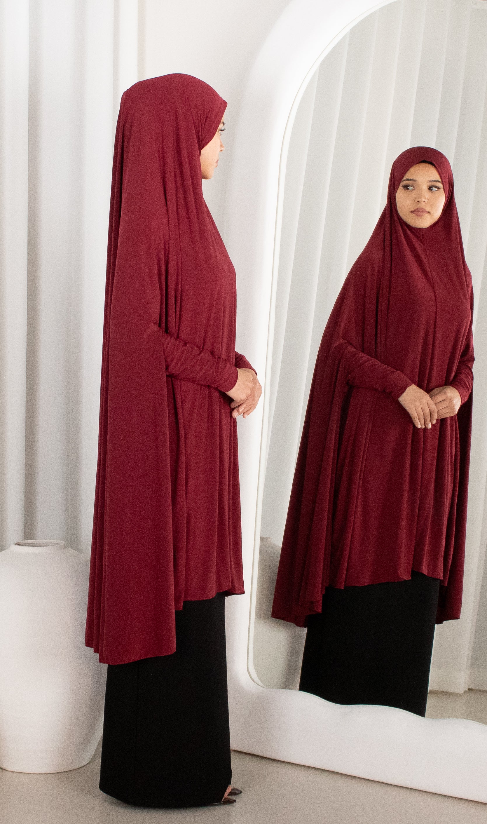 Woman wearing a burgundy hijab and black outfit standing in front of a mirror.