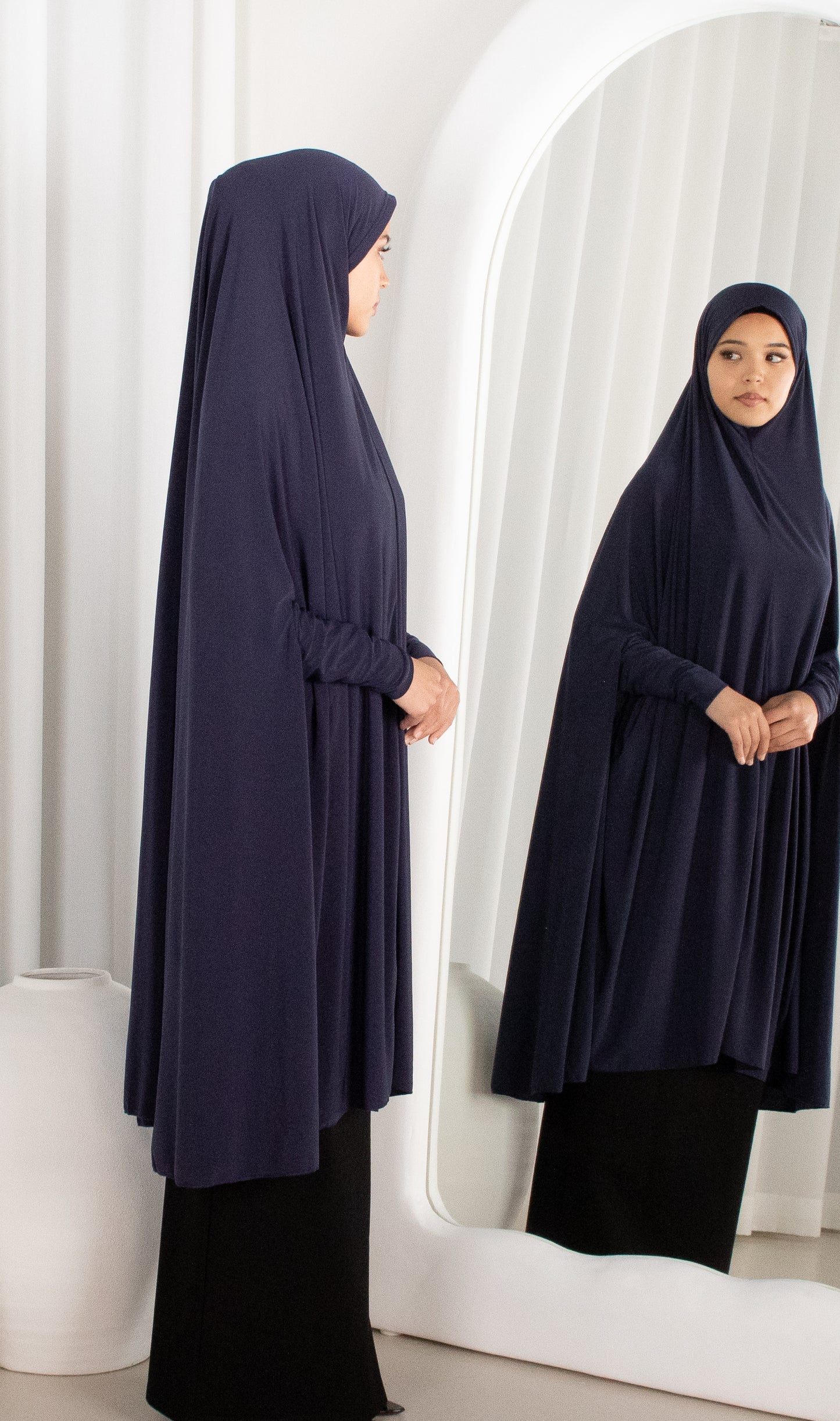 Woman wearing a navy blue hijab and dress standing in front of a mirror.