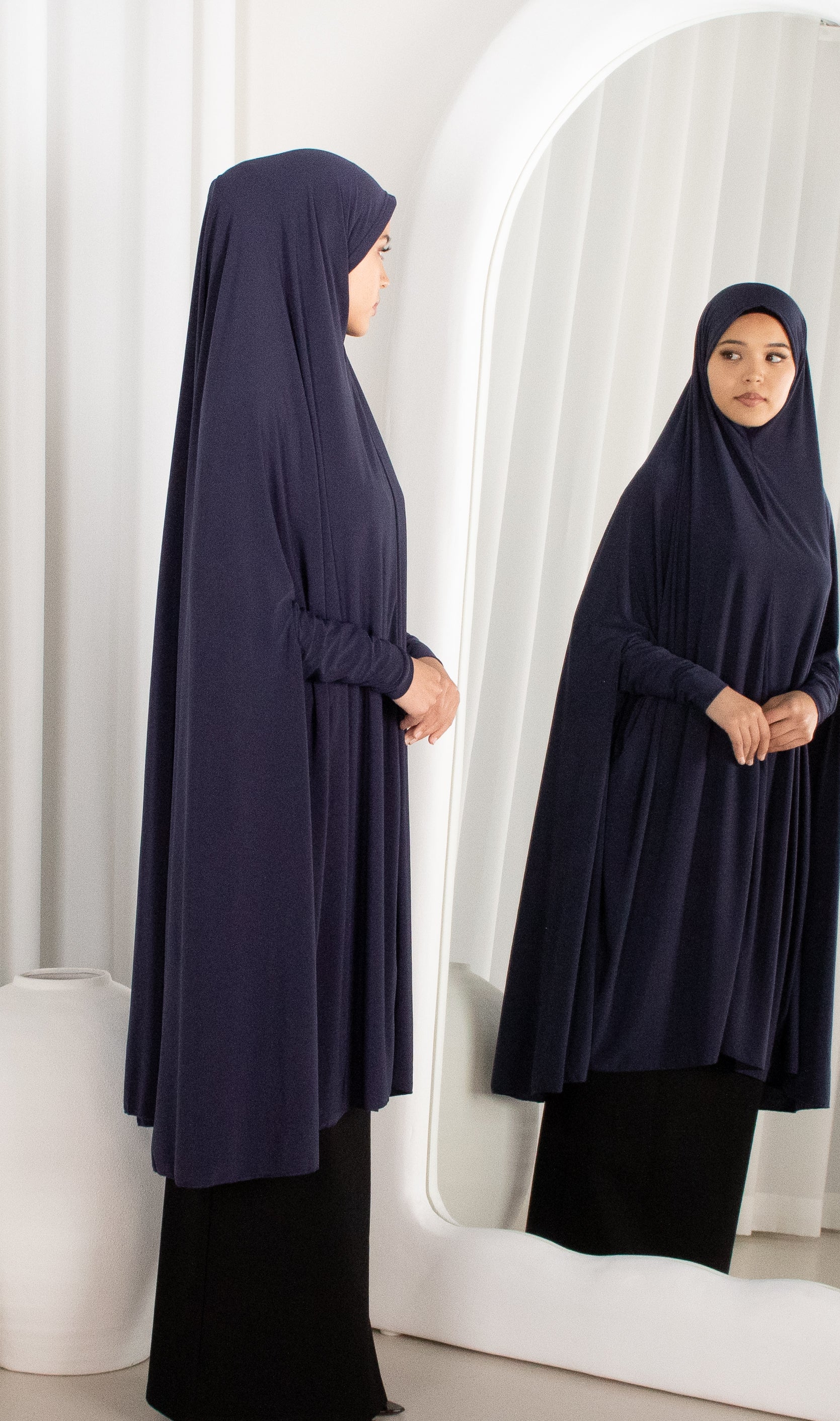 Woman wearing a navy blue hijab and dress standing in front of a mirror.