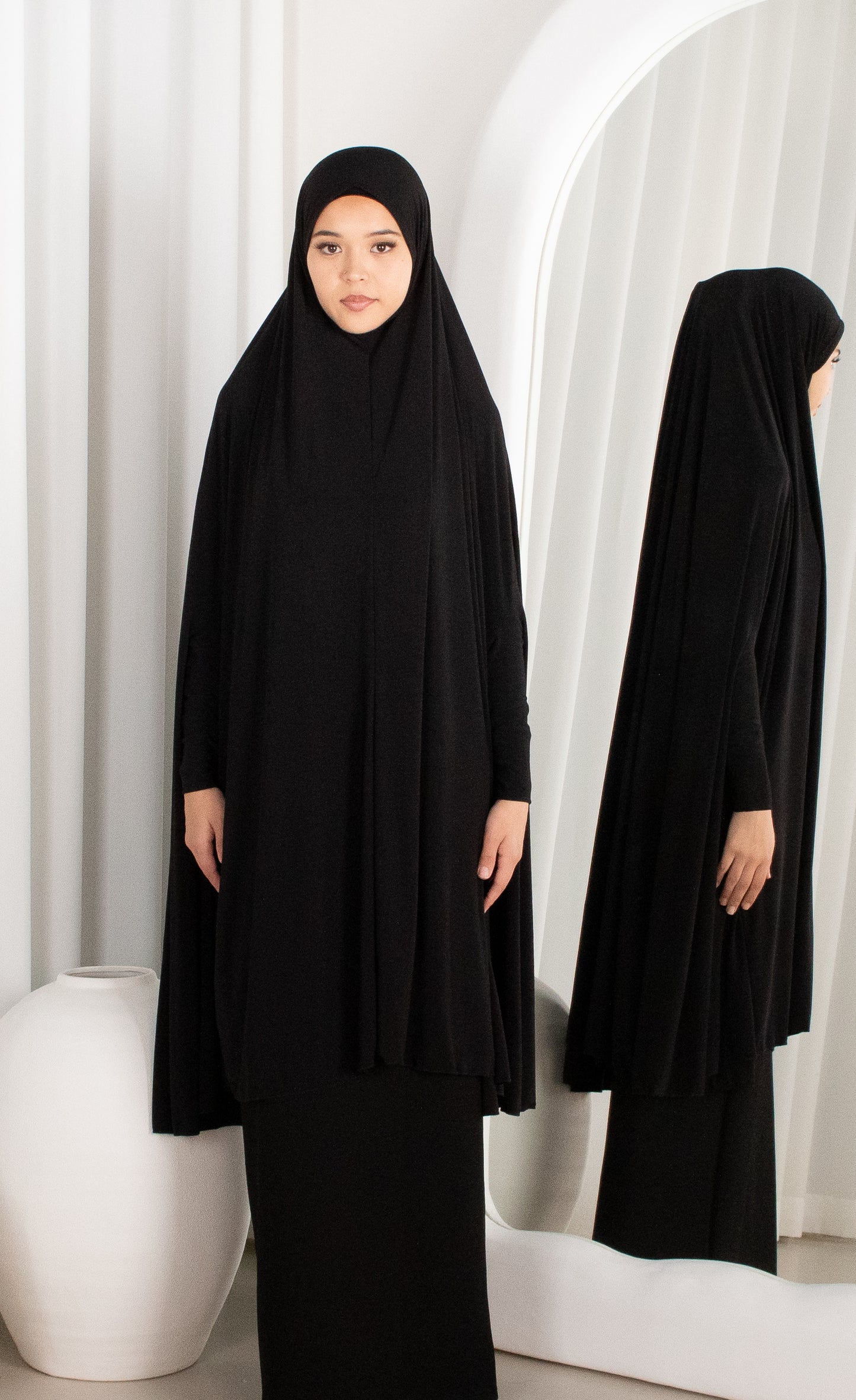 Woman wearing a black abaya in front of a white curtain