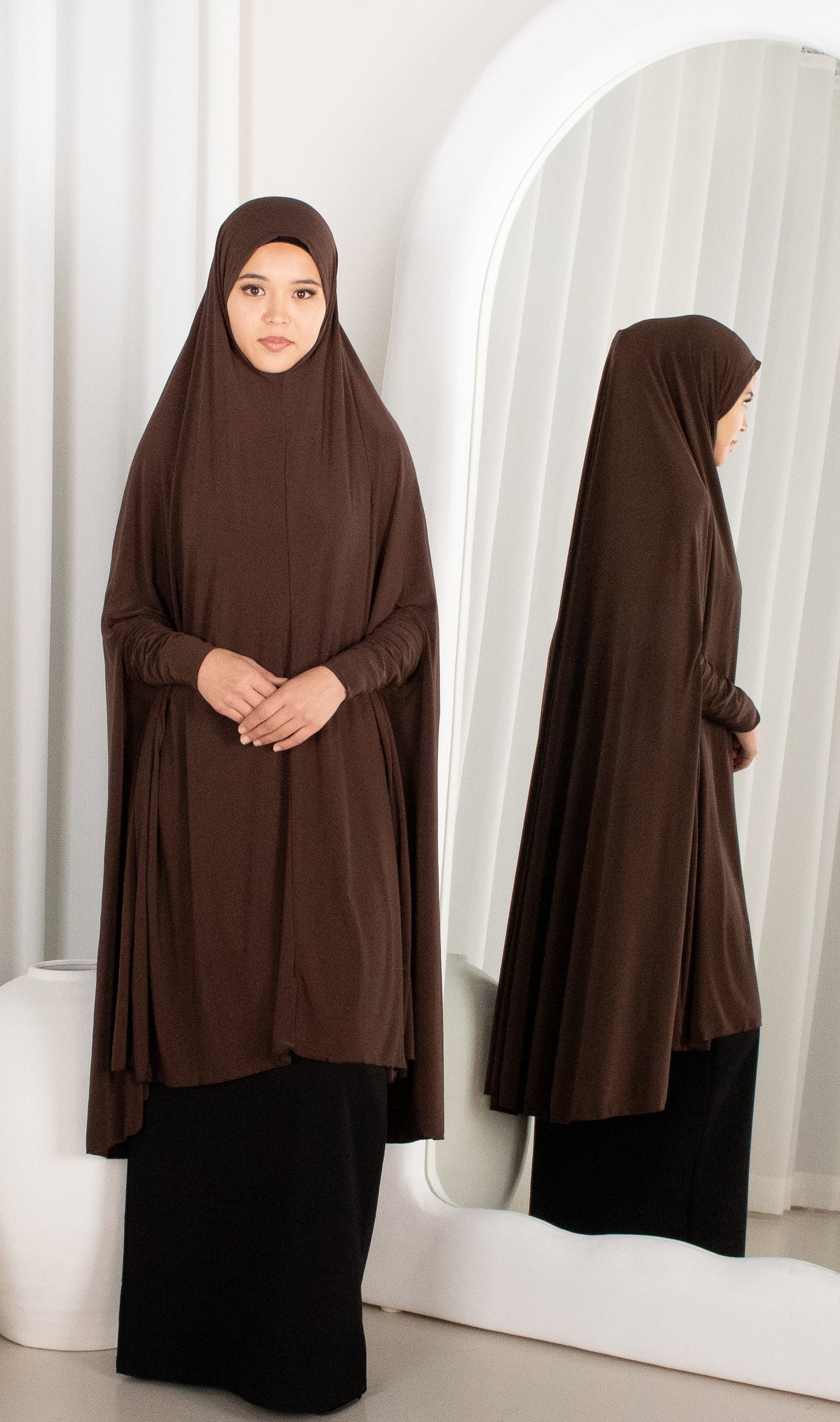Woman wearing a brown hijab and black outfit standing in front of a mirror.