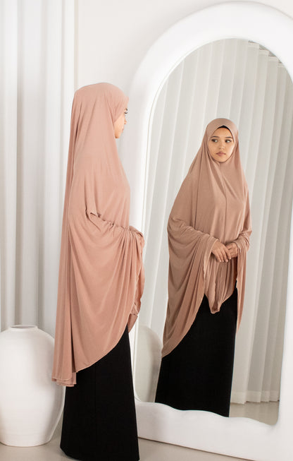 Woman wearing a beige hijab and black dress standing in front of a mirror.