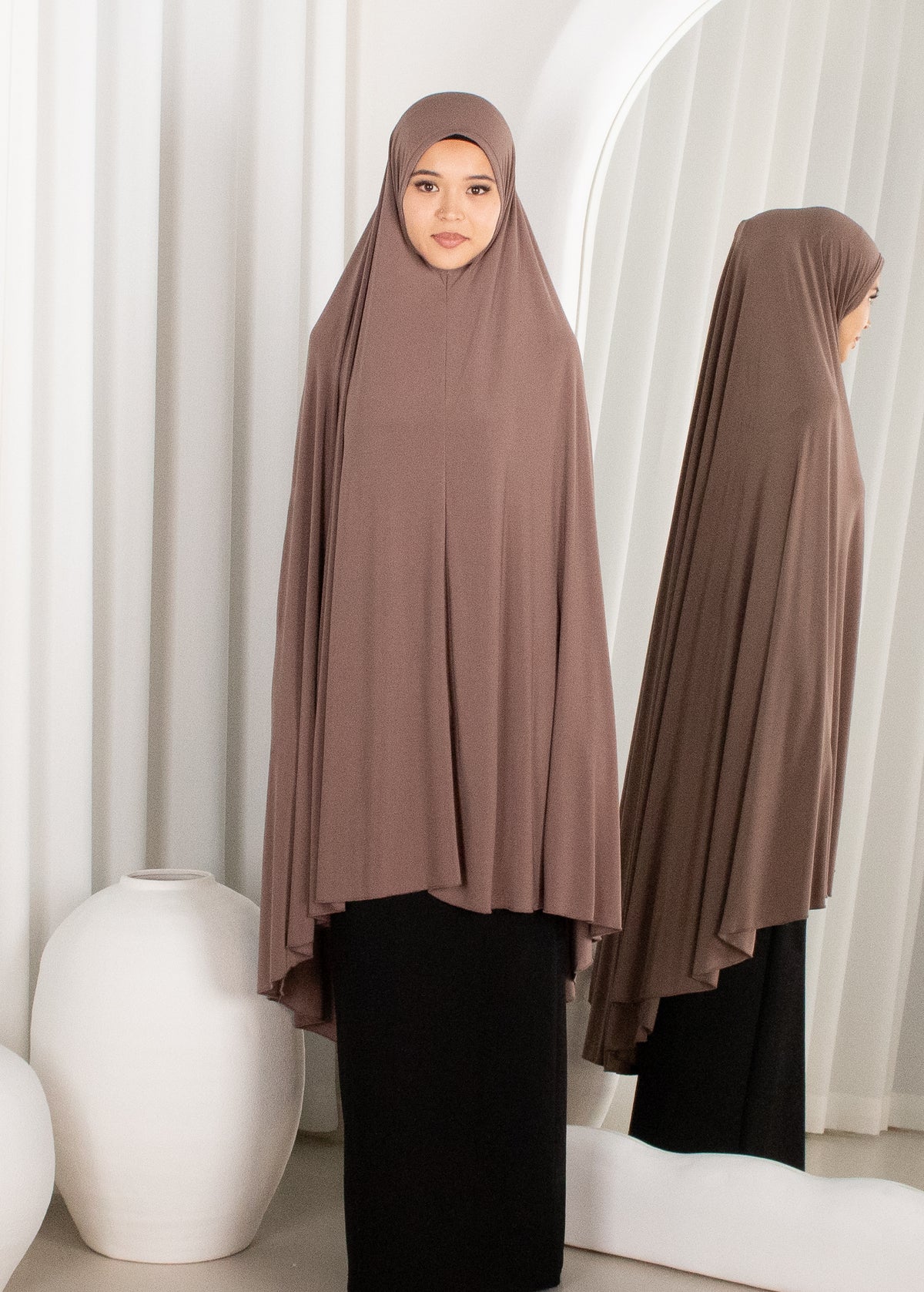 Woman wearing a brown hijab and black outfit in front of white curtains