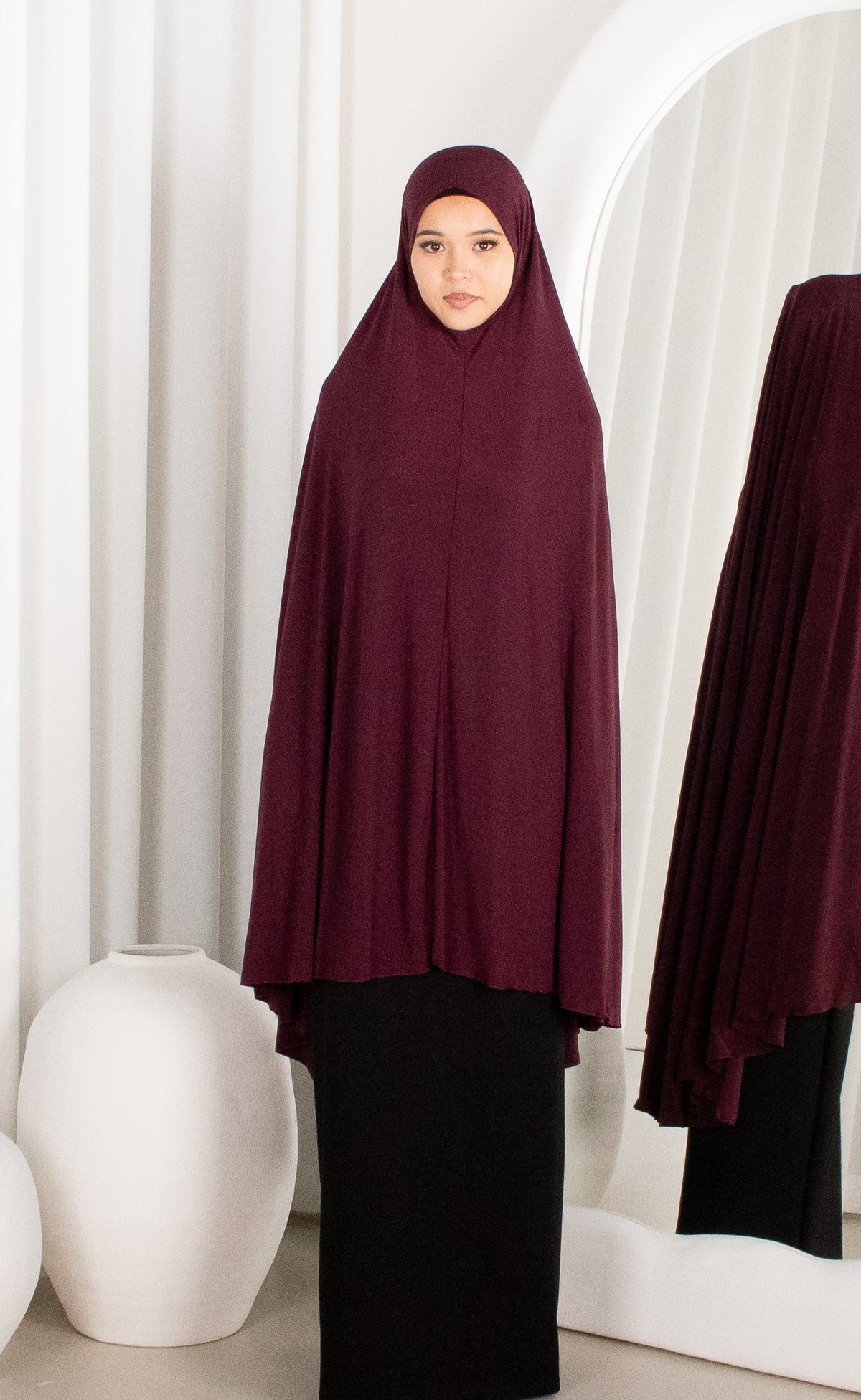 Woman wearing a burgundy hijab and black outfit standing in a room with white curtains.