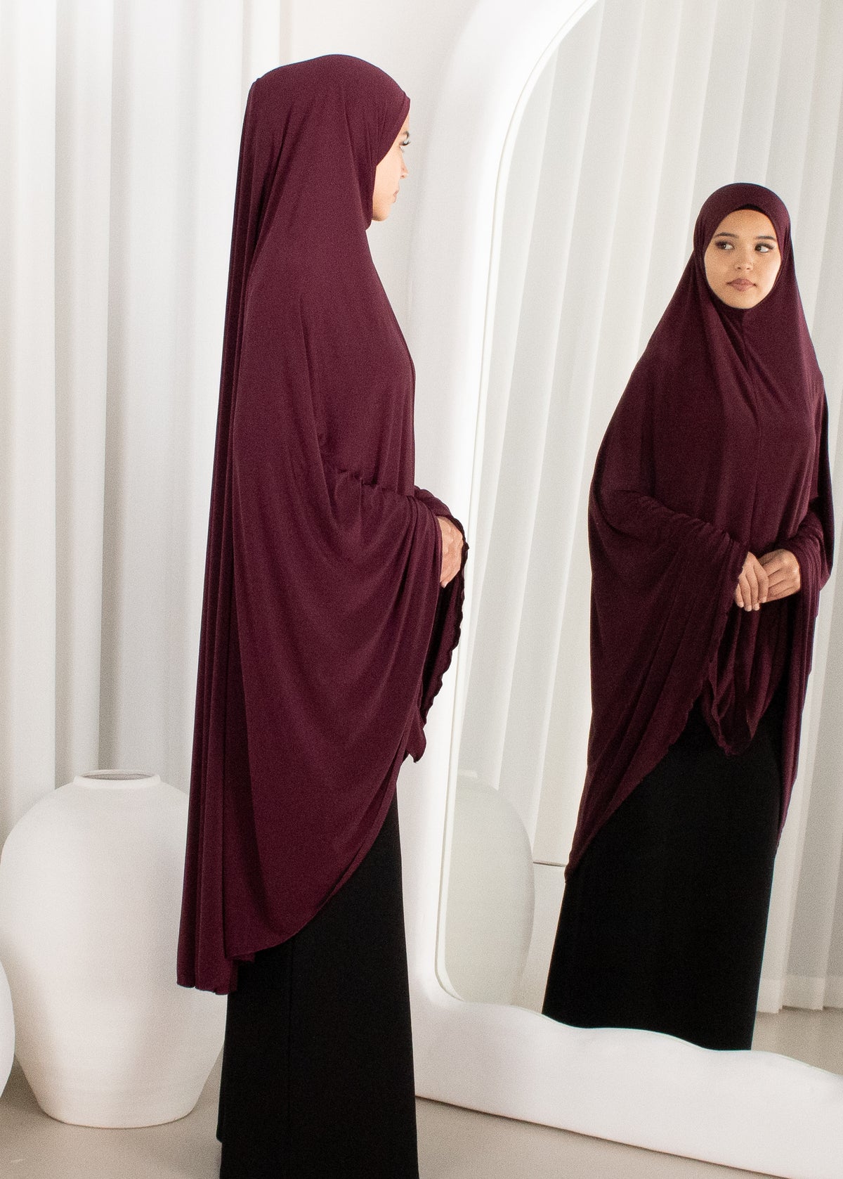 Woman wearing a burgundy hijab and black dress standing in front of a mirror.