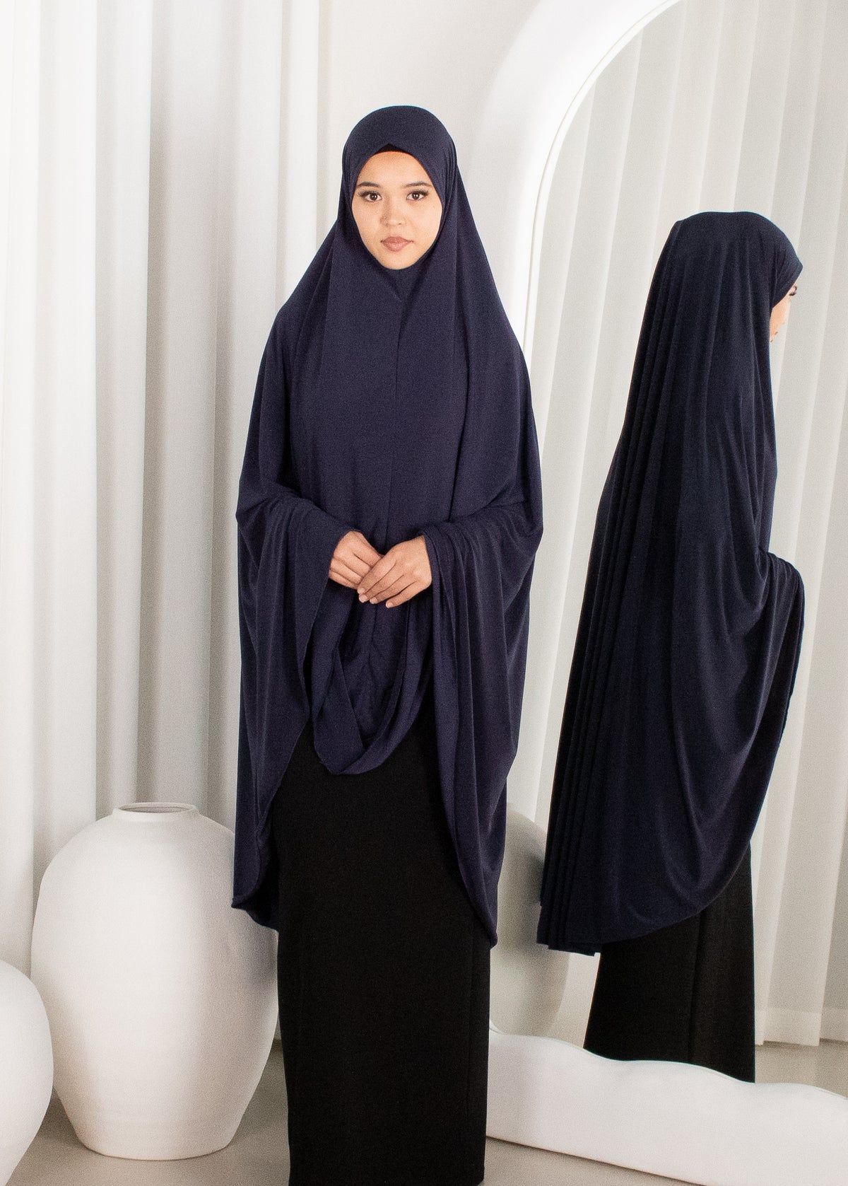 Woman wearing a navy blue hijab and black dress standing in front of a white curtain.