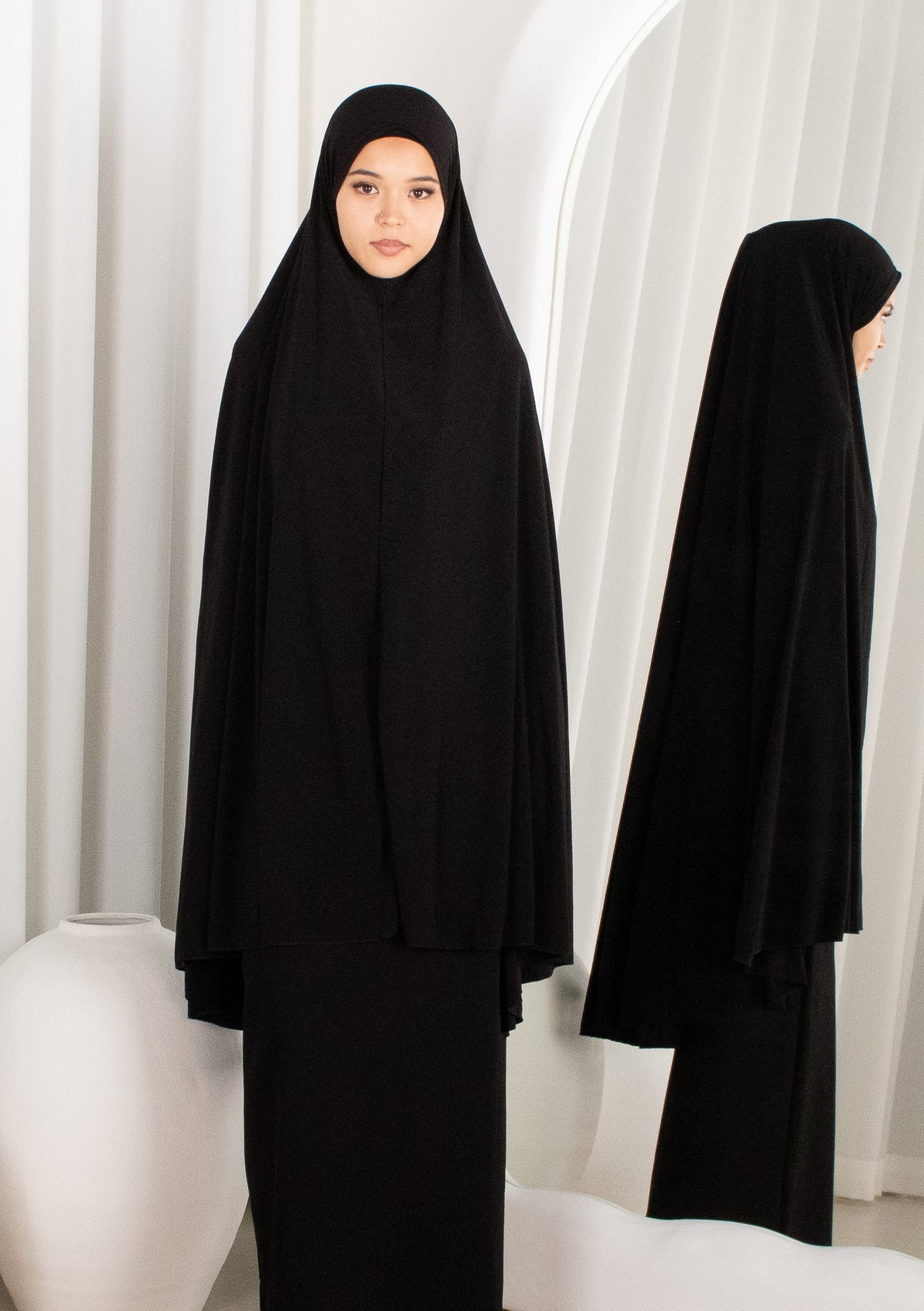 Black abaya worn by a model against a white curtain background