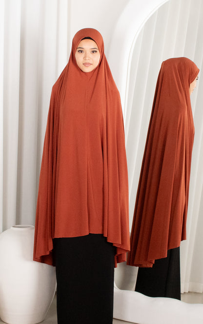 Person wearing a rust-colored hijab with a white curtain background