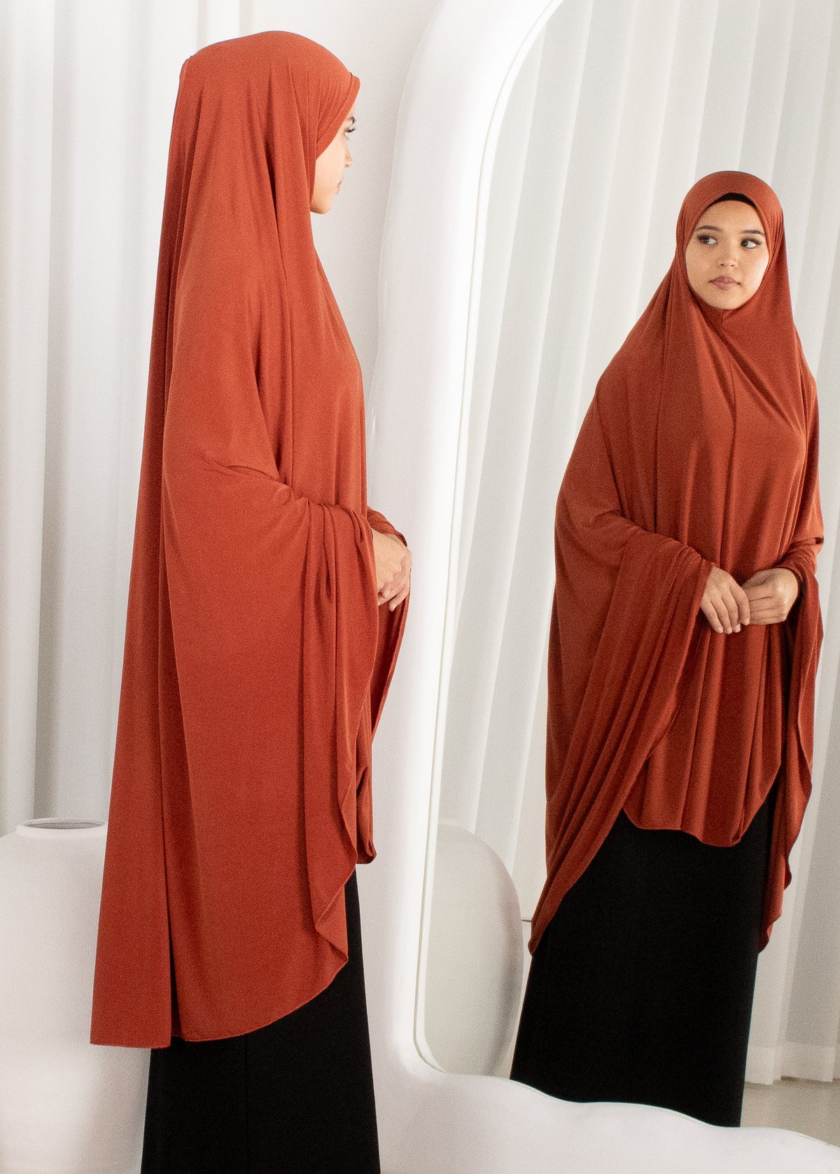Person wearing a rust-colored hijab and black outfit standing against a white curtain.