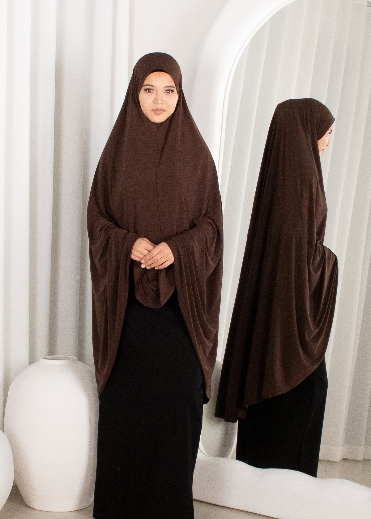 Woman wearing a brown hijab and black dress standing in front of white curtains.