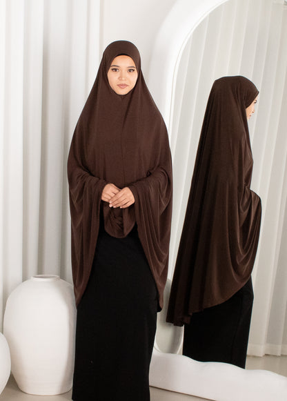 Woman wearing a brown hijab and black dress standing in front of white curtains.