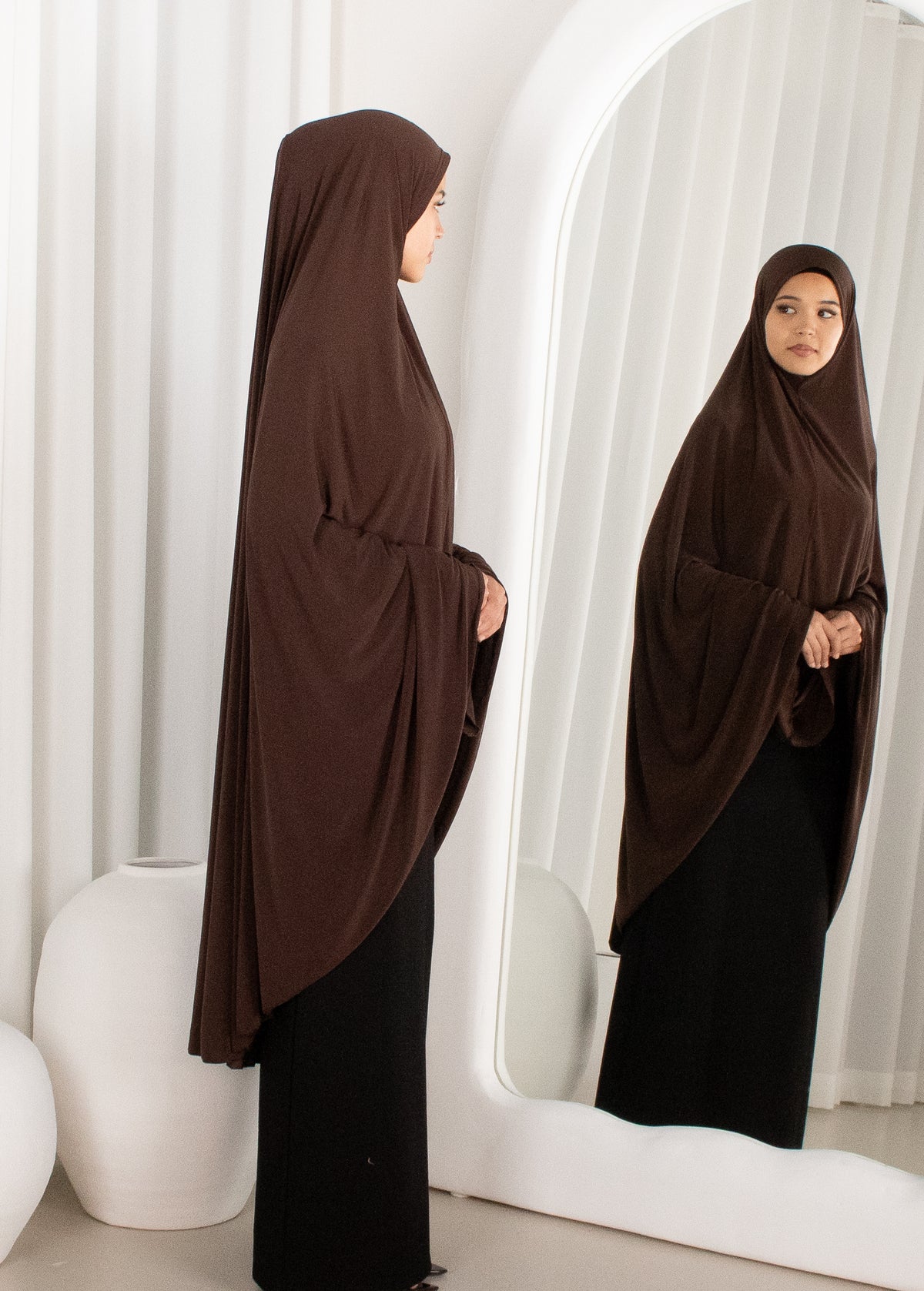Woman wearing a brown hijab and black dress standing in front of a mirror.