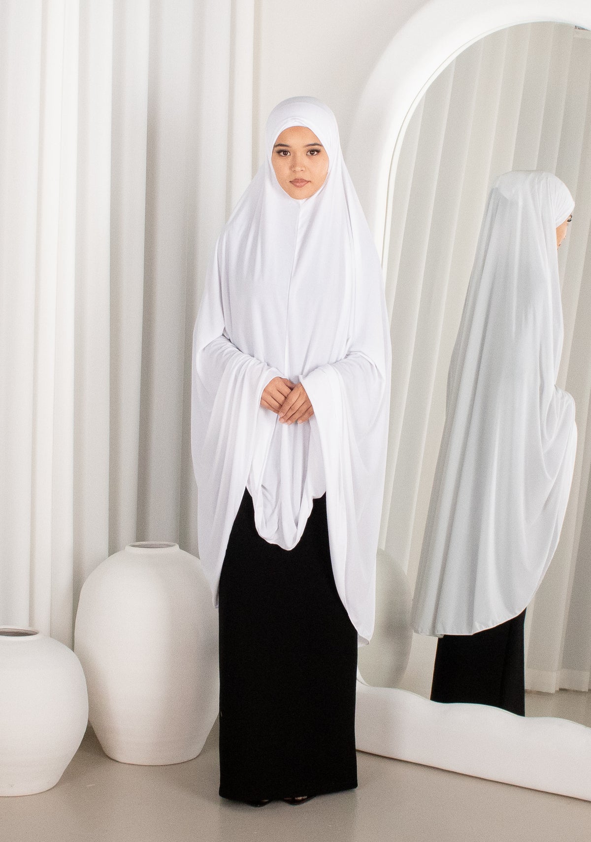 Woman wearing a white hijab and black outfit in a minimalistic room.