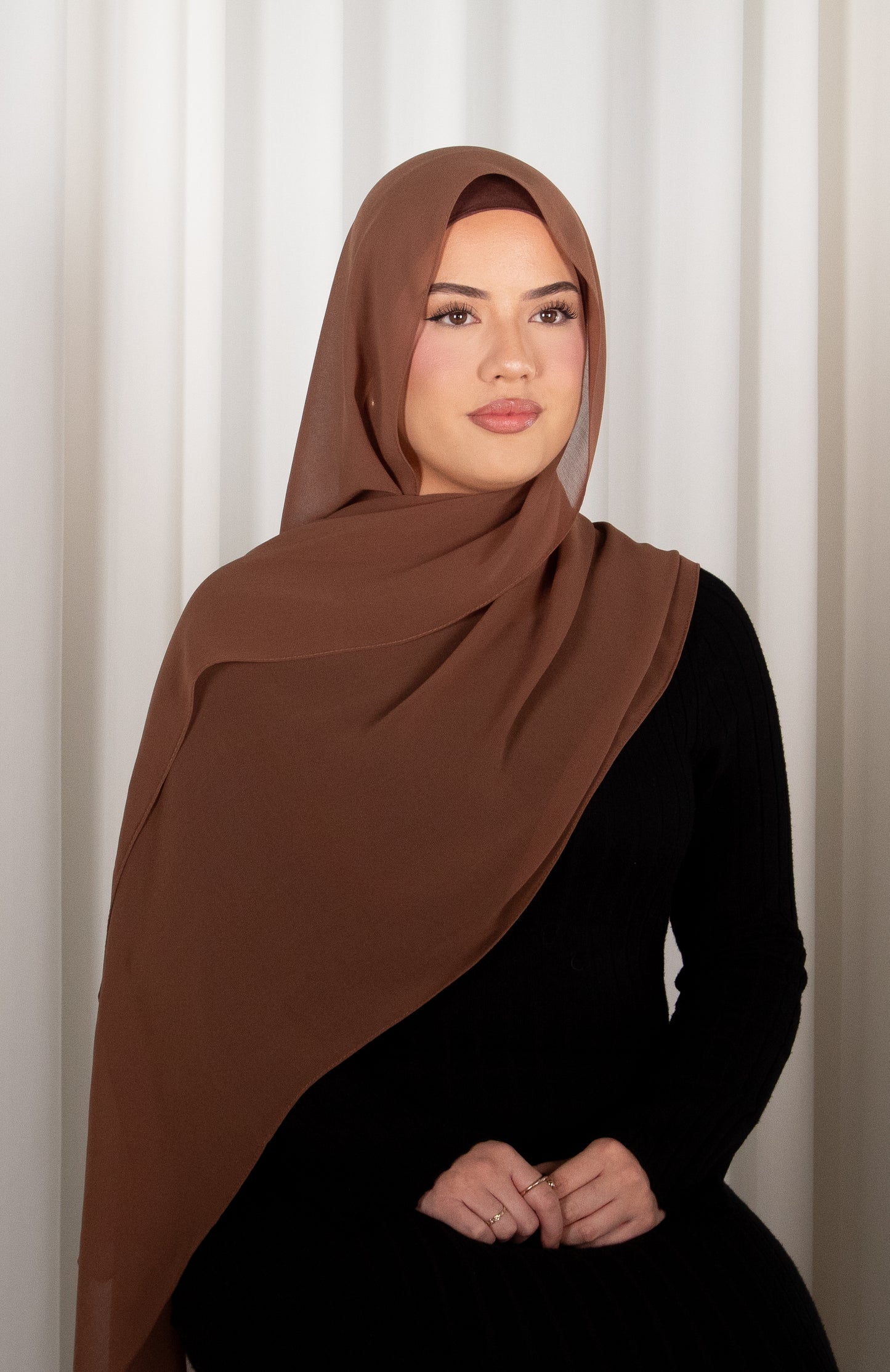 Woman wearing a brown hijab and black outfit against a white curtain background