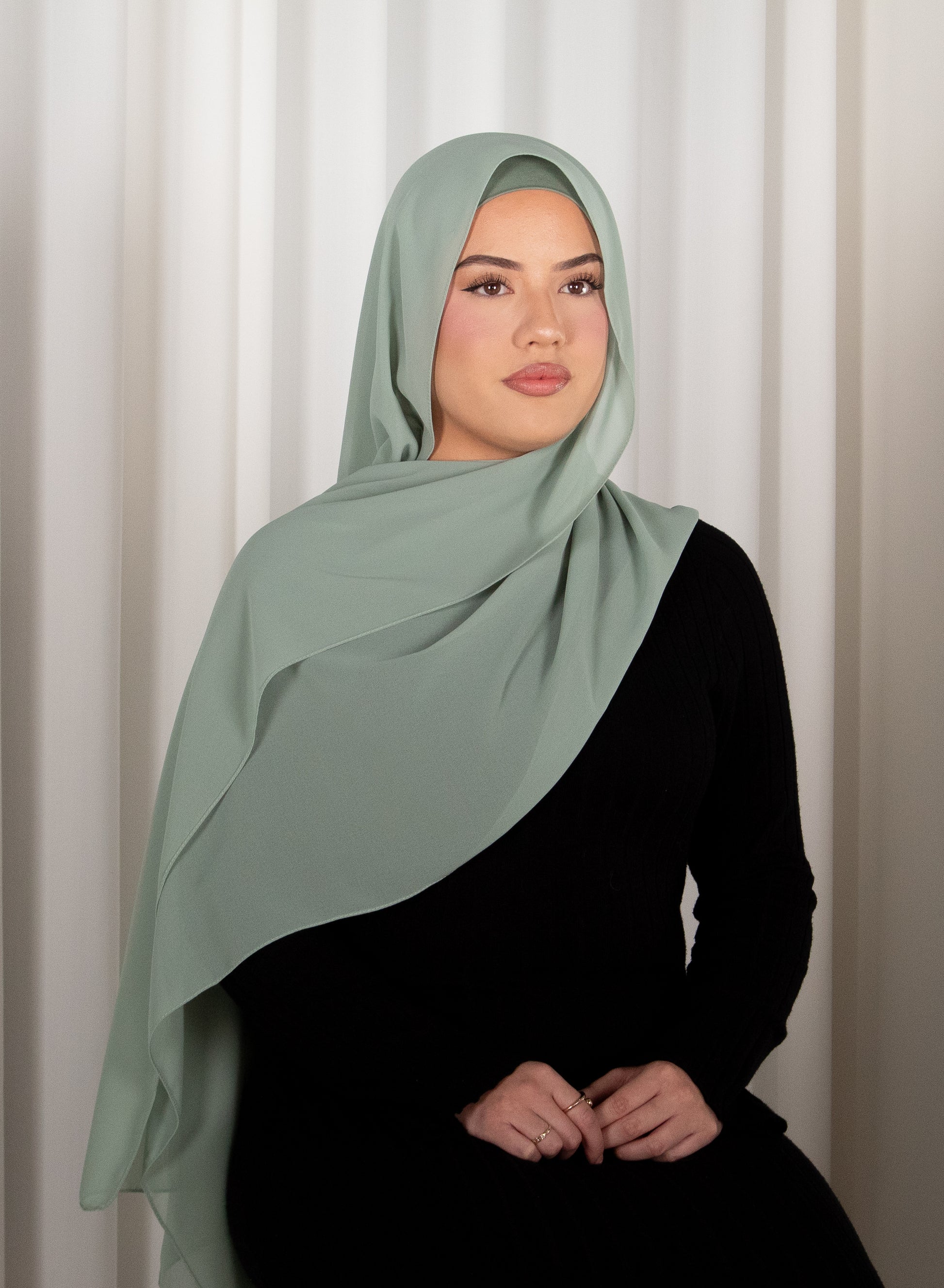 Woman wearing a light green hijab and black outfit against a white curtain background