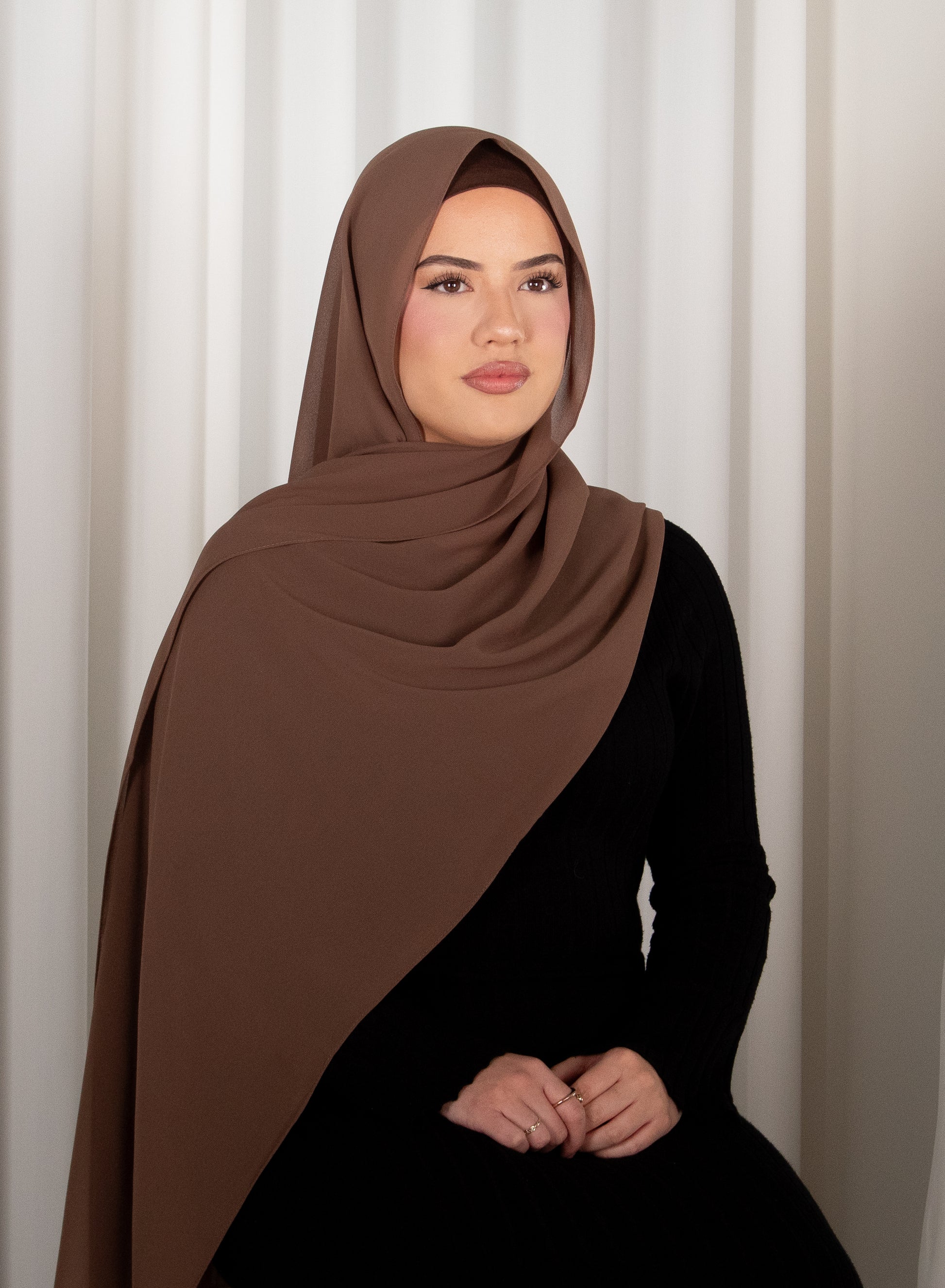 Woman wearing a brown hijab and black outfit against a white curtain background