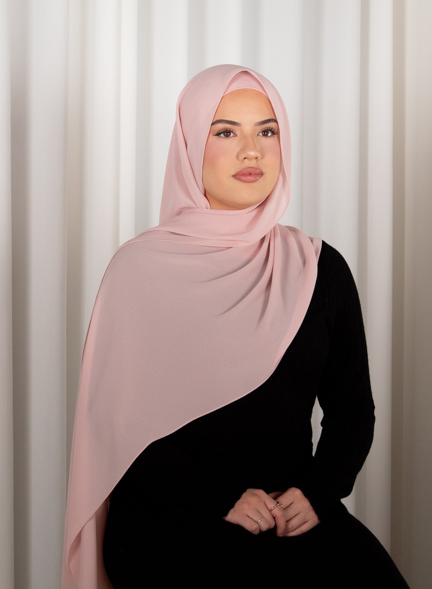Woman wearing a pink hijab and black outfit against a white curtain background