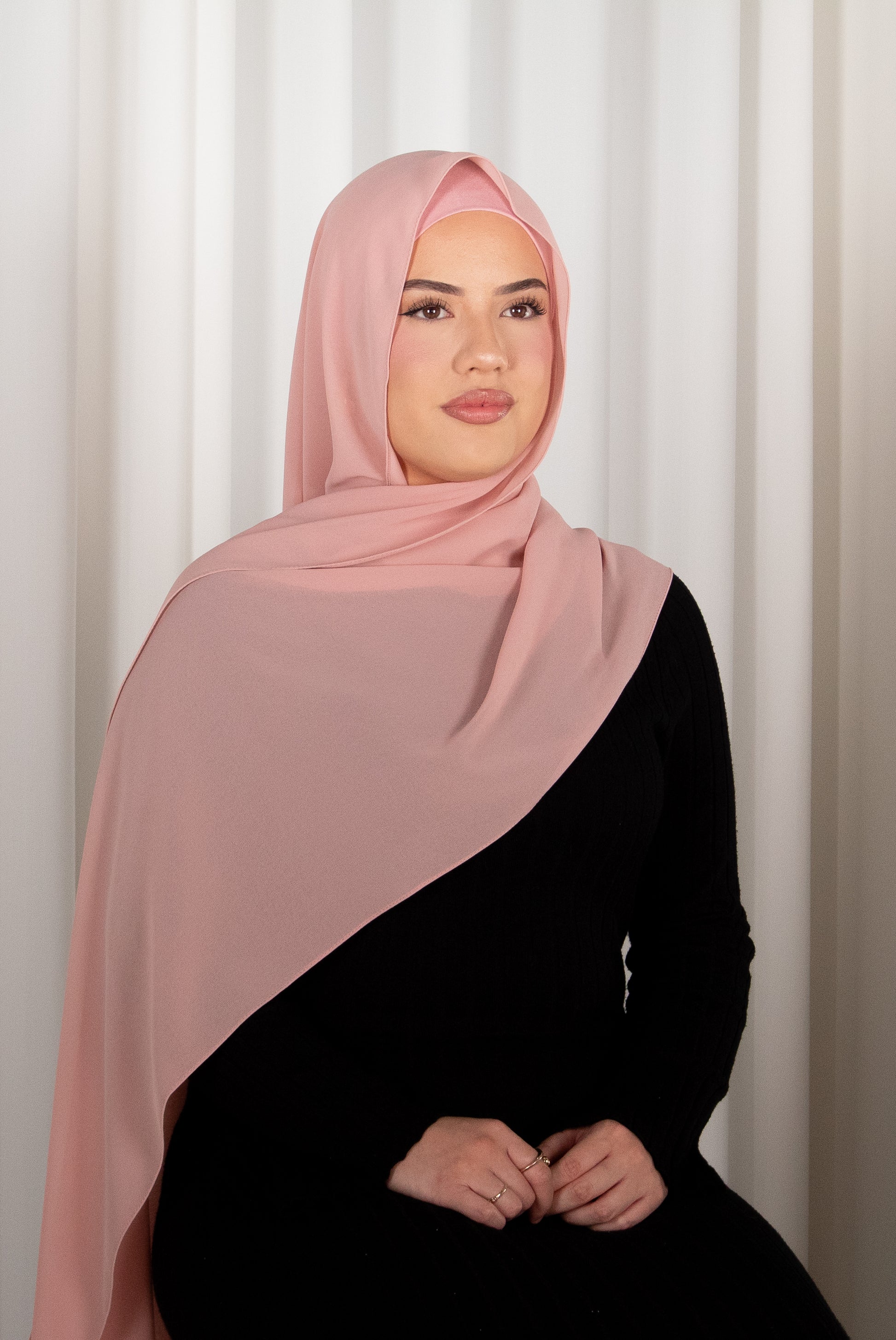 Woman wearing a pink hijab and black outfit against a white curtain background