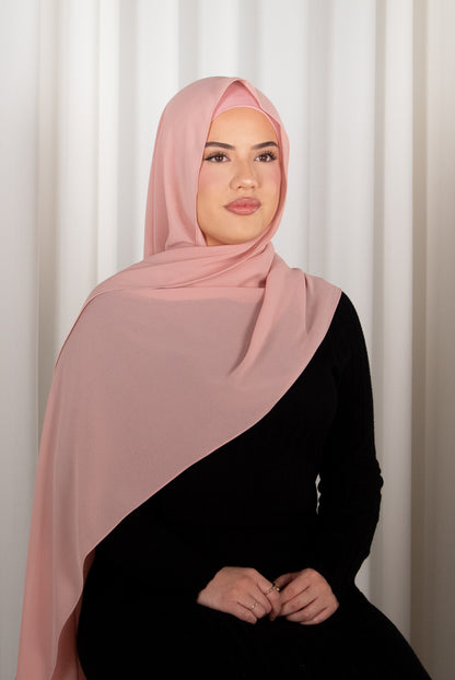 Woman wearing a pink hijab and black outfit against a white curtain background