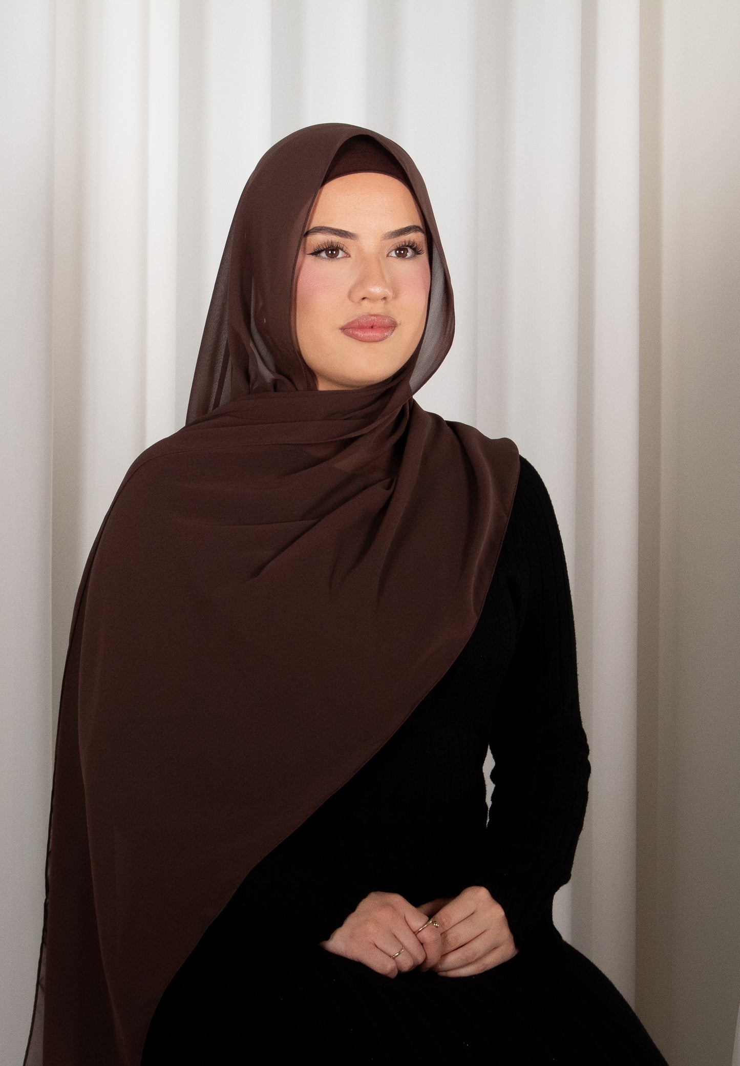 Woman wearing a brown hijab and black outfit against a white curtain background