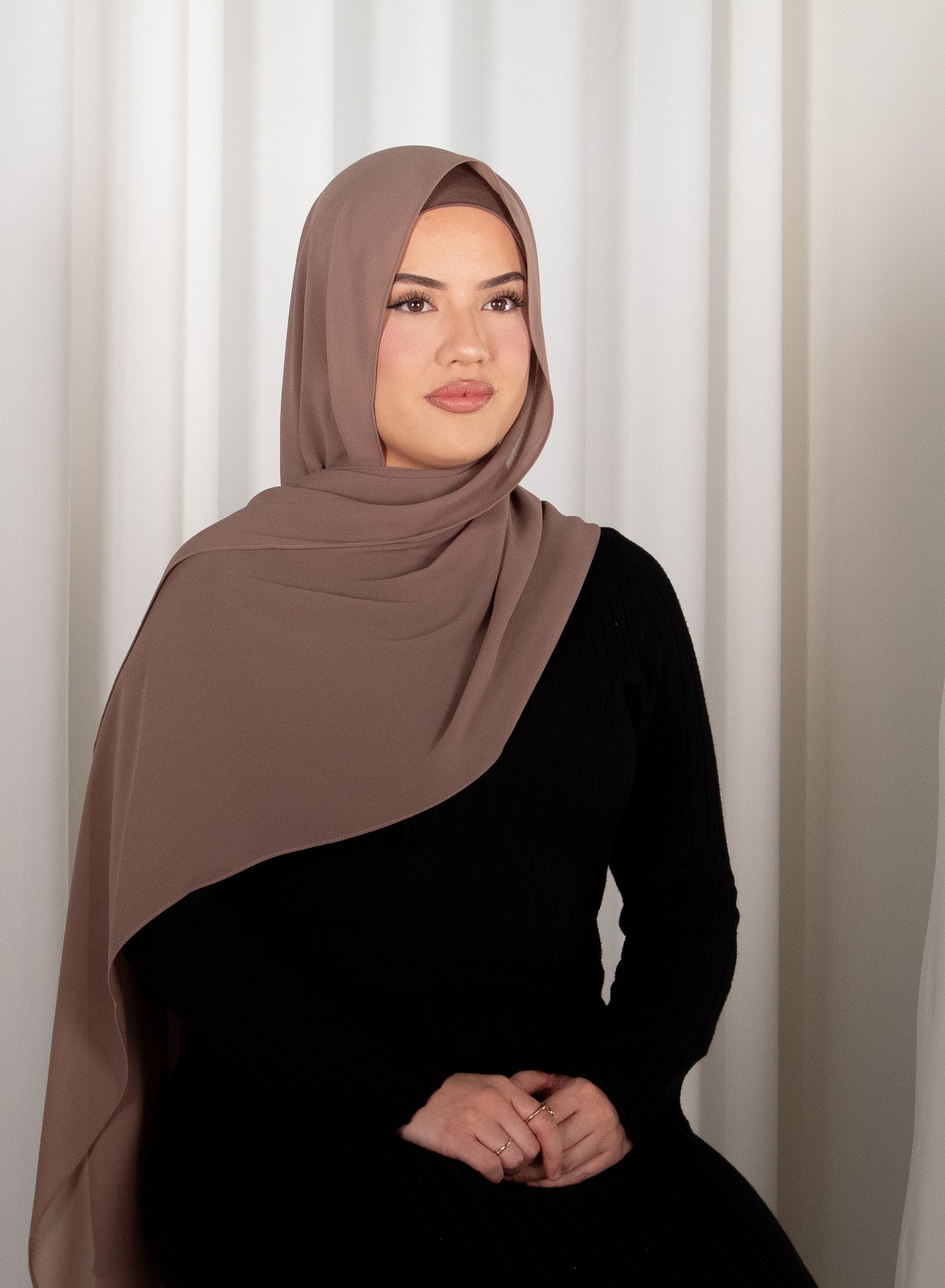 Woman wearing a brown hijab and black outfit against a white curtain background