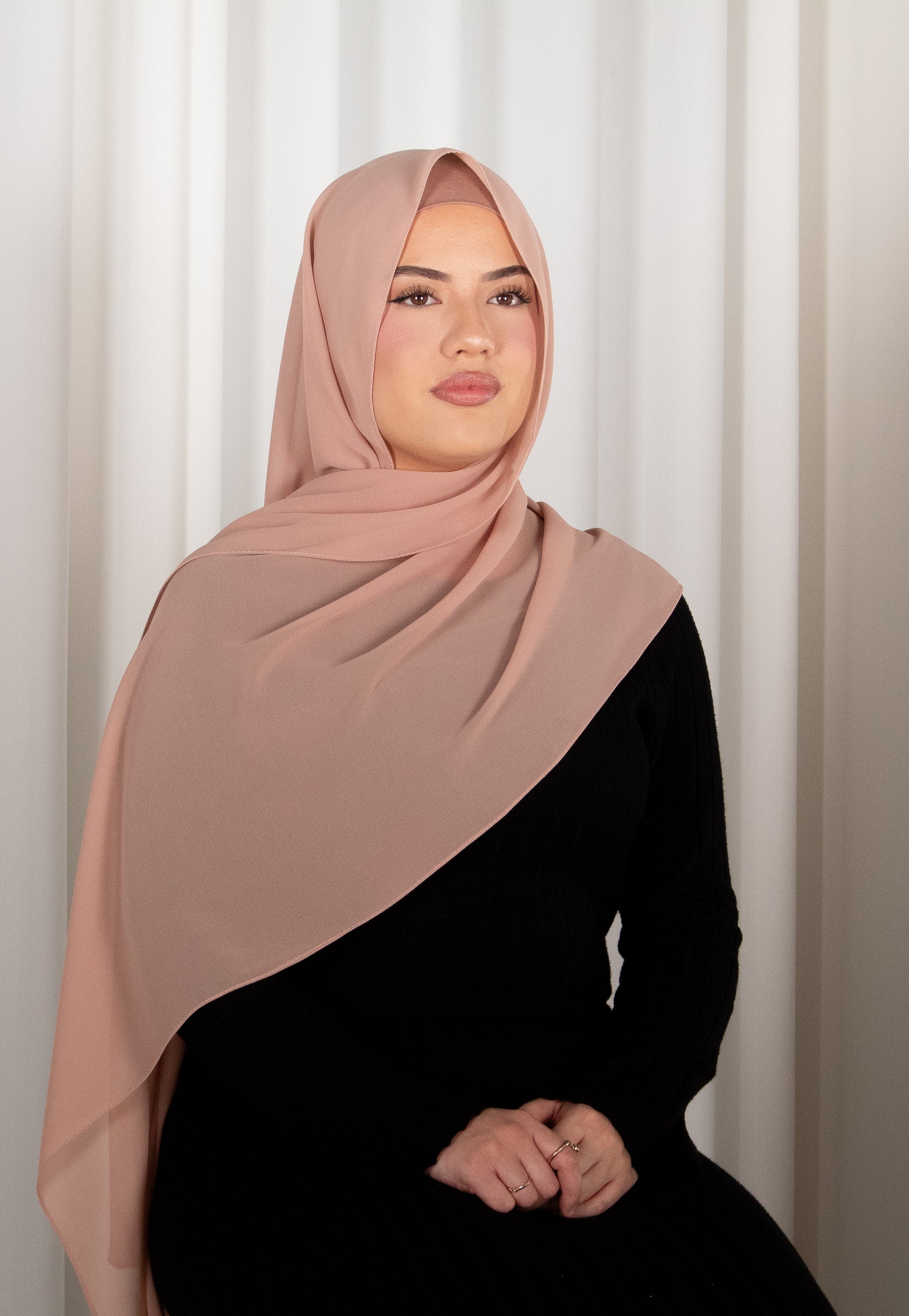 Woman wearing a pink hijab and black outfit against a white curtain background