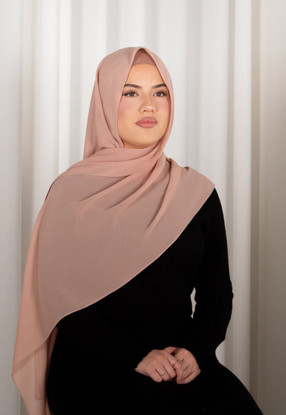 Woman wearing a pink hijab and black outfit against a white curtain background