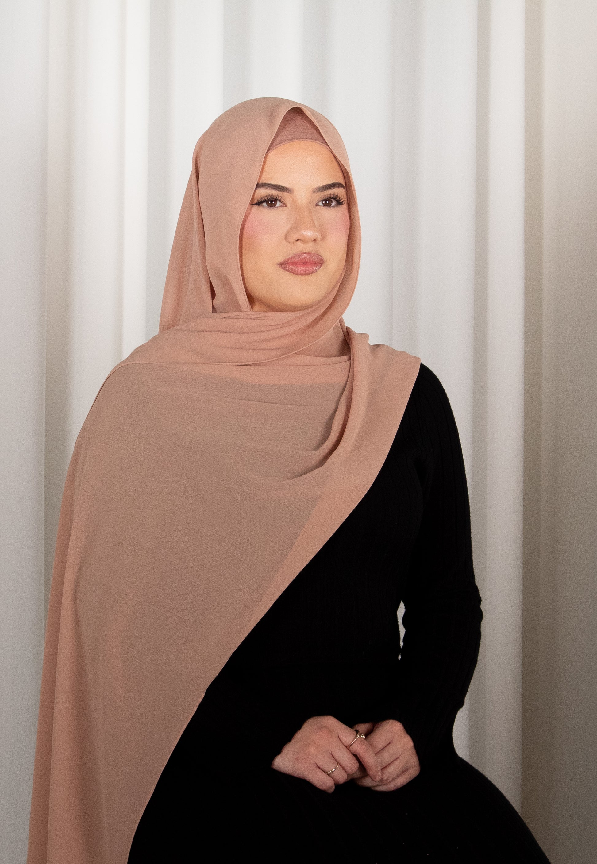 Woman wearing a brown hijab and black outfit against a white curtain background