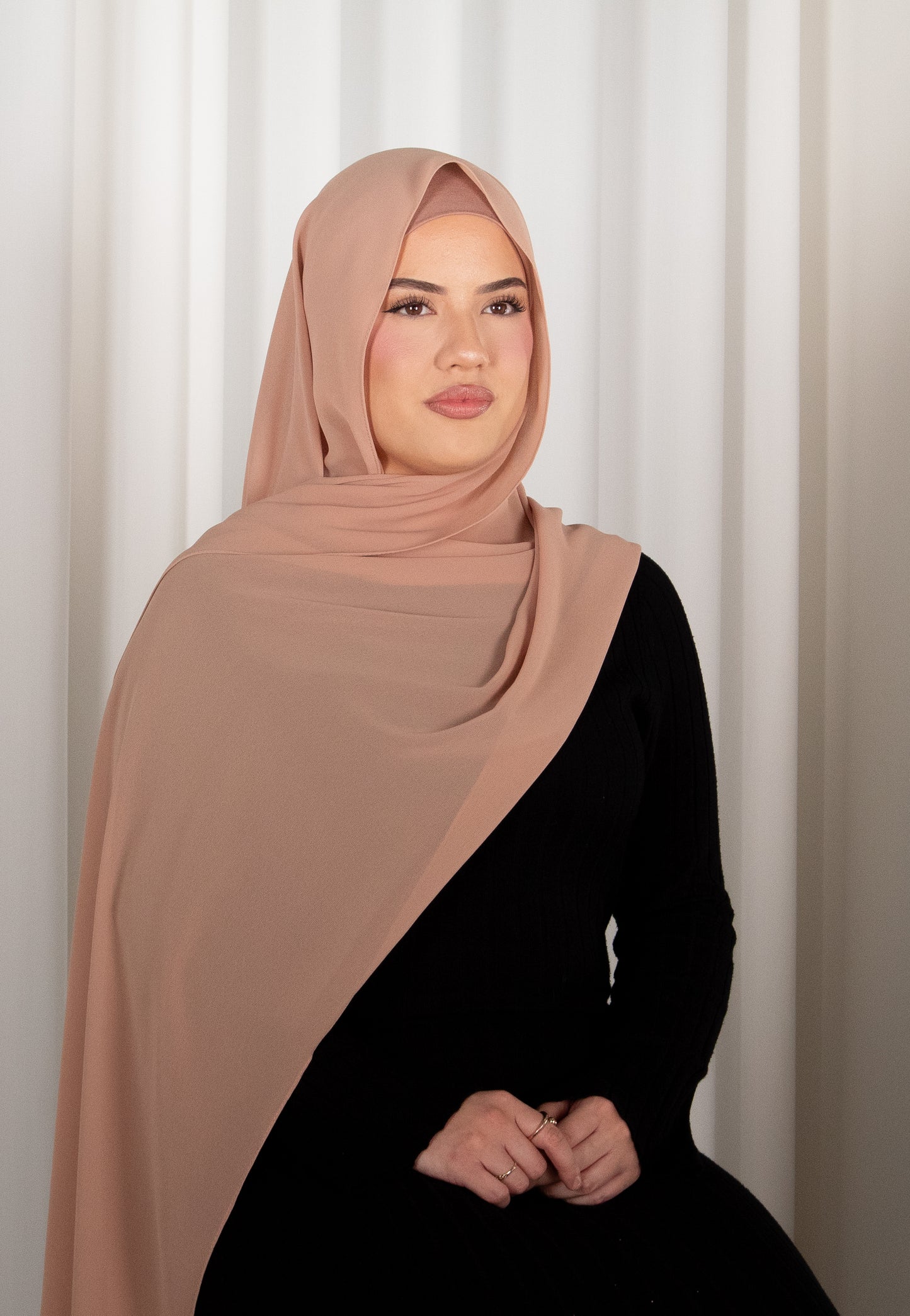 Woman wearing a brown hijab and black outfit against a white curtain background