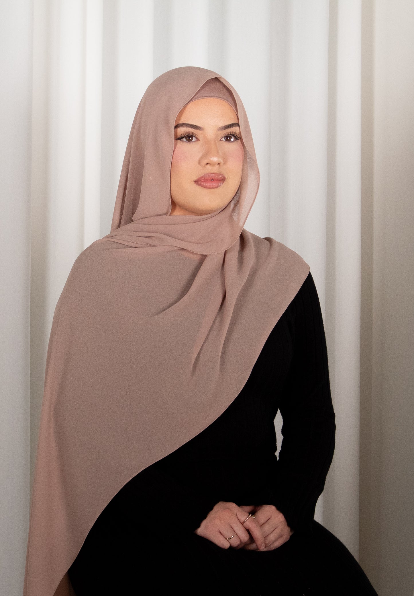 Woman wearing a beige hijab and black outfit against a white curtain background