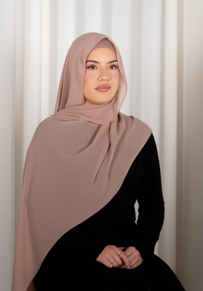 Woman wearing a beige hijab and black outfit against a white curtain background