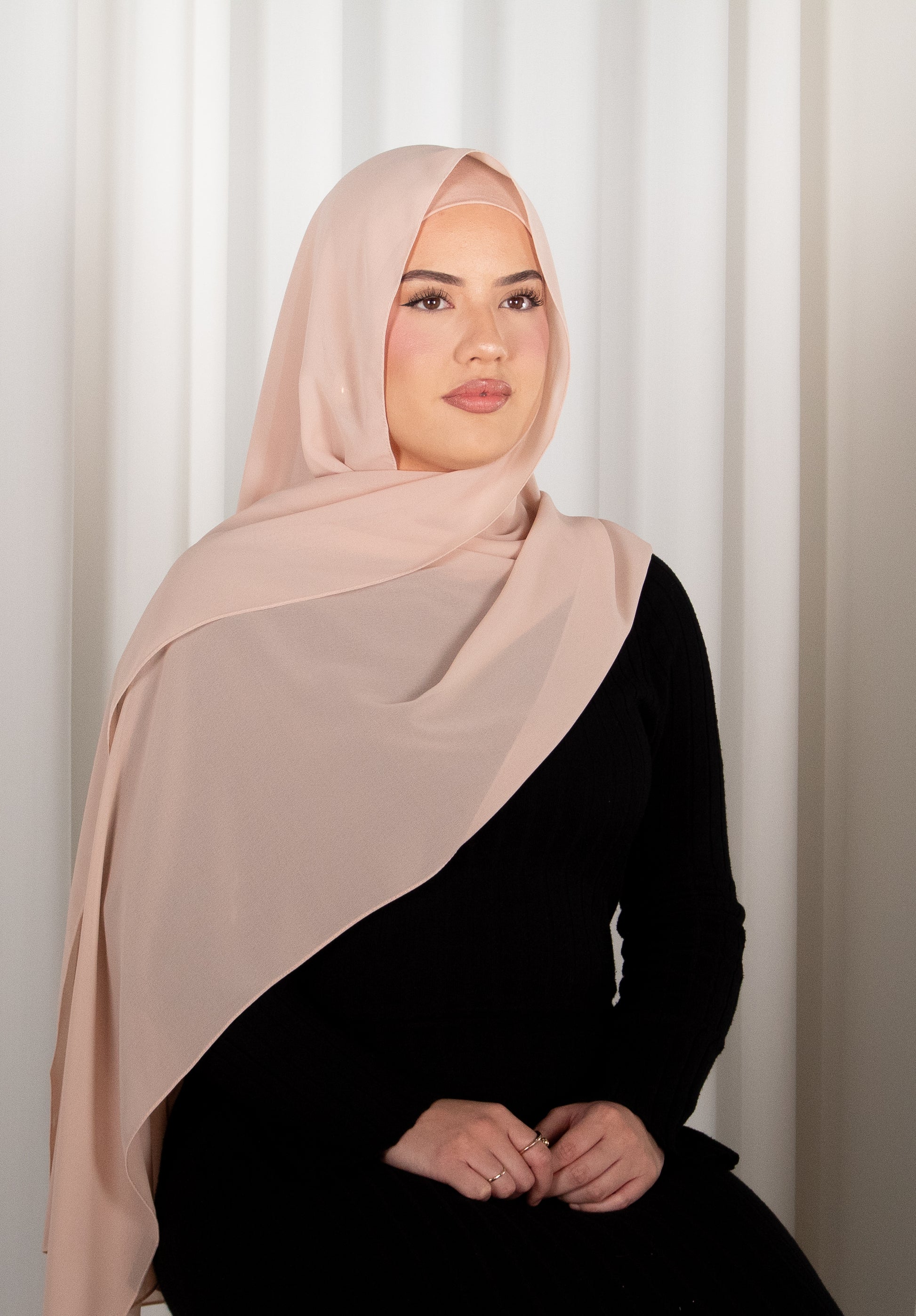 Woman wearing a beige hijab and black outfit against a white curtain background