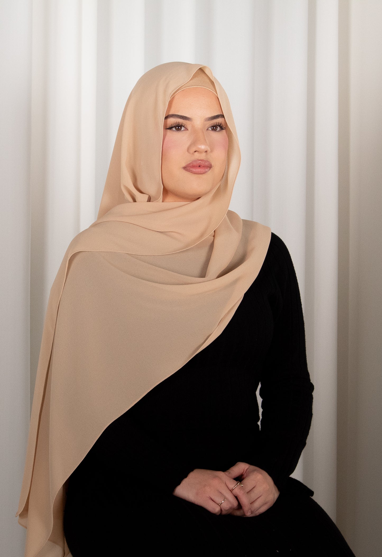 Woman wearing a beige hijab and black outfit against a white curtain background