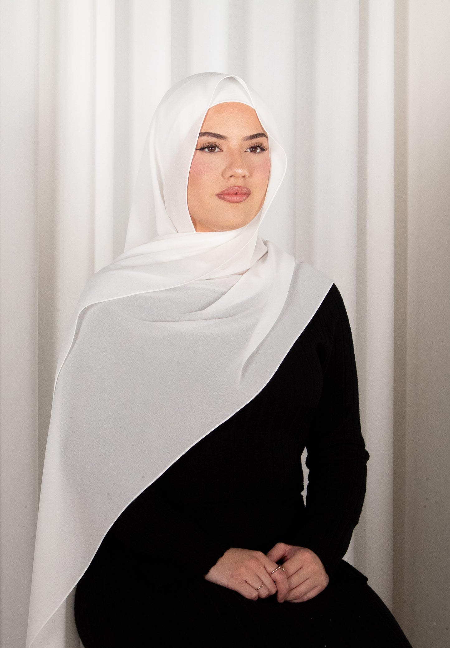 Woman wearing a white hijab and black outfit against a white curtain background