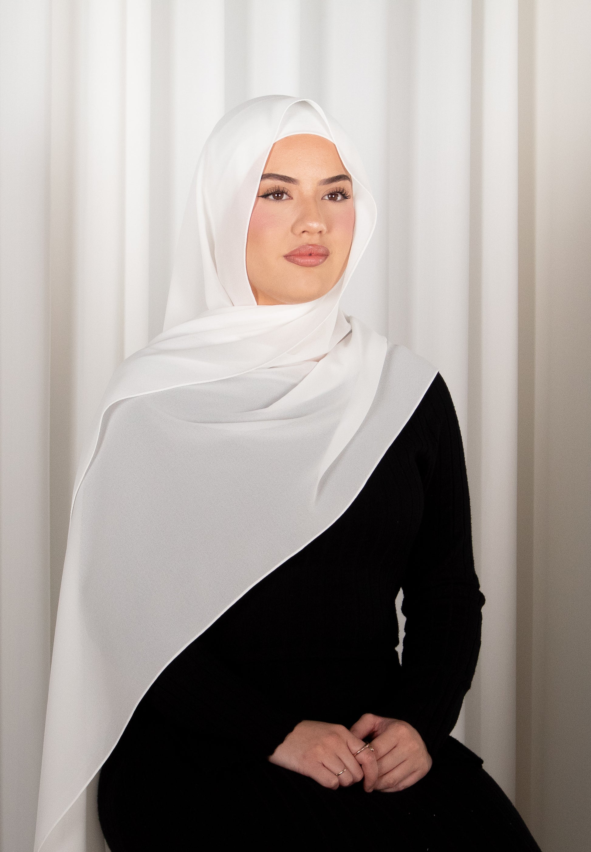 Woman wearing a white hijab and black outfit against a white curtain background