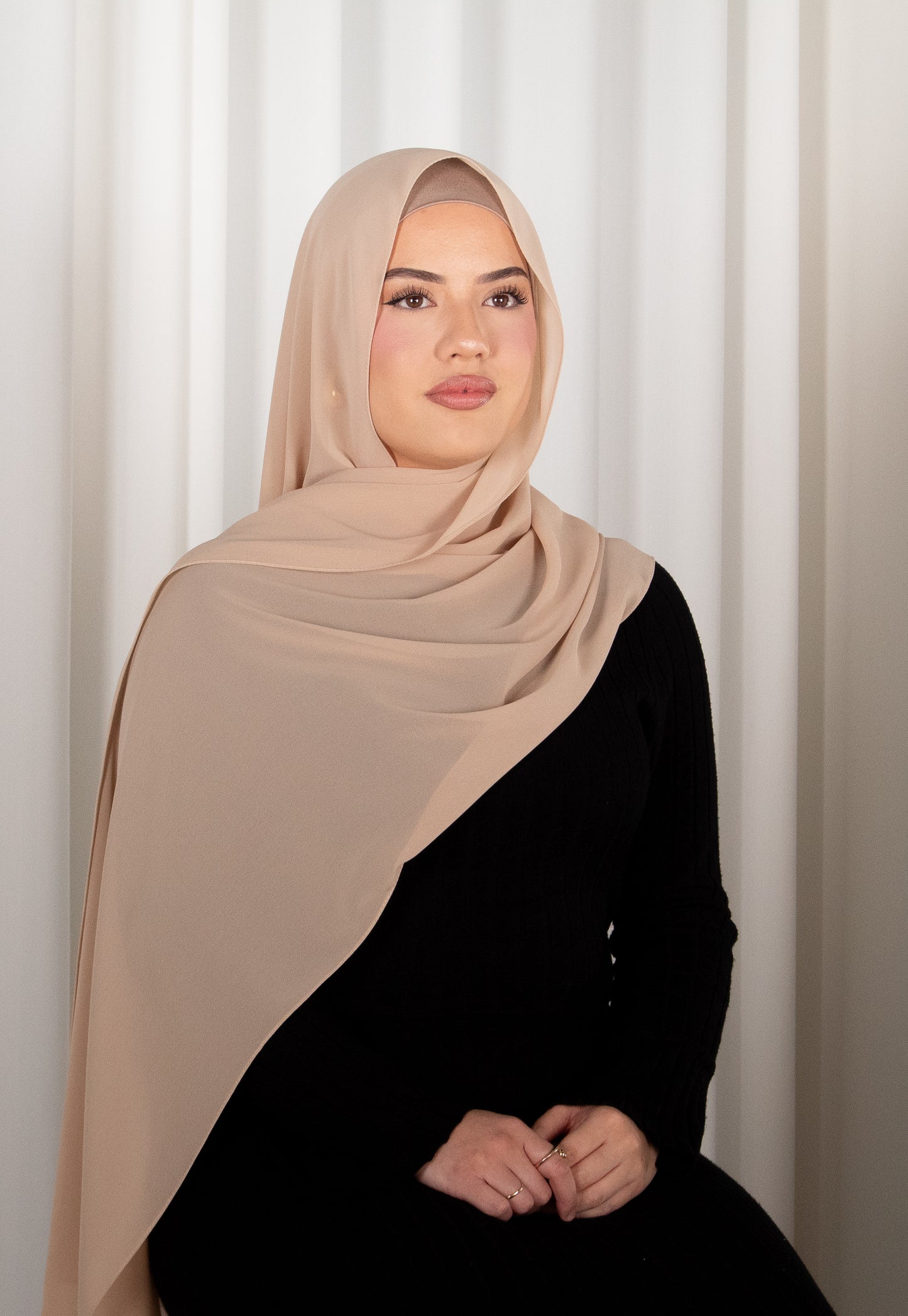 Woman wearing a beige hijab against a white curtain background