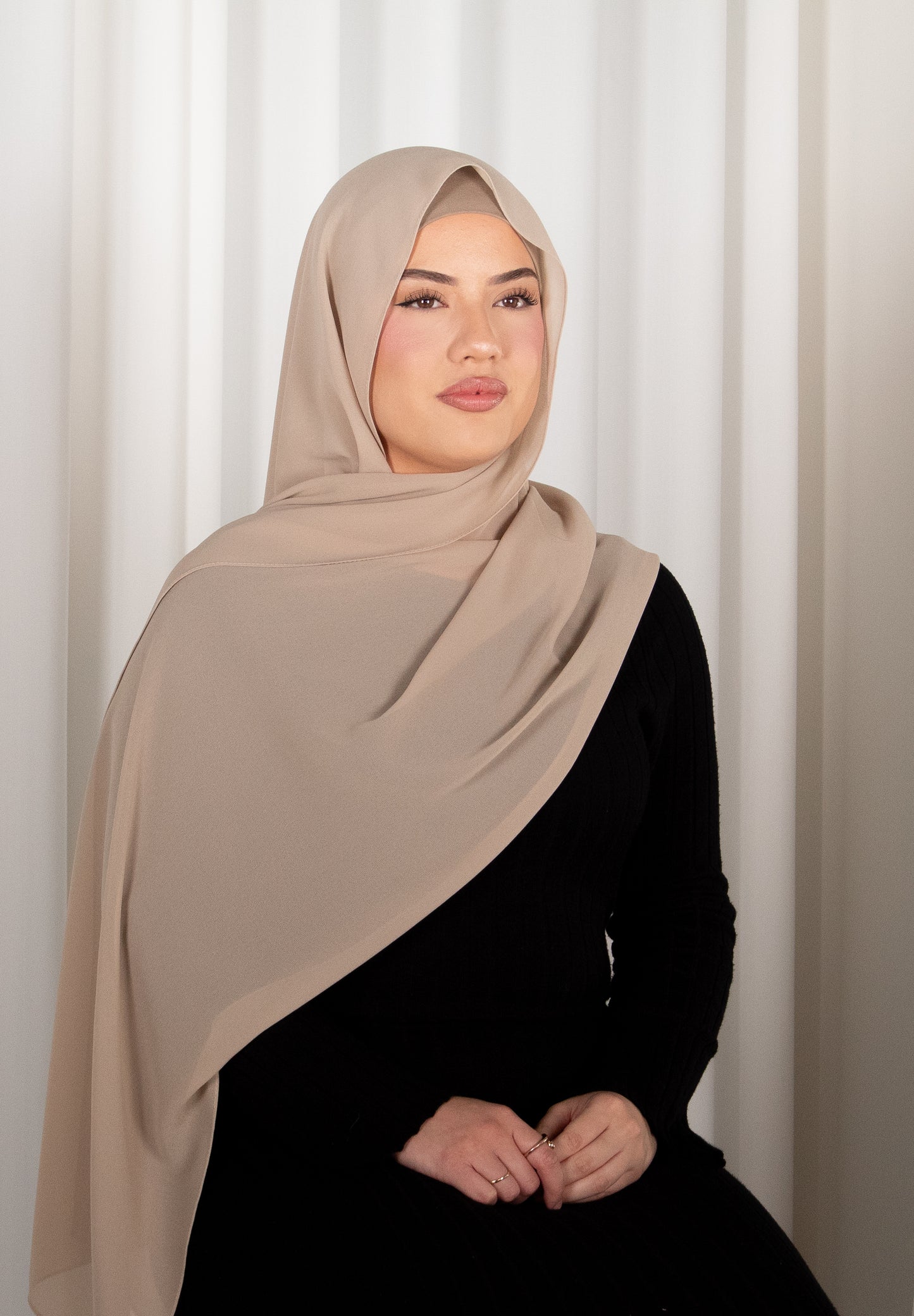 Woman wearing a beige hijab and black outfit against a white curtain background