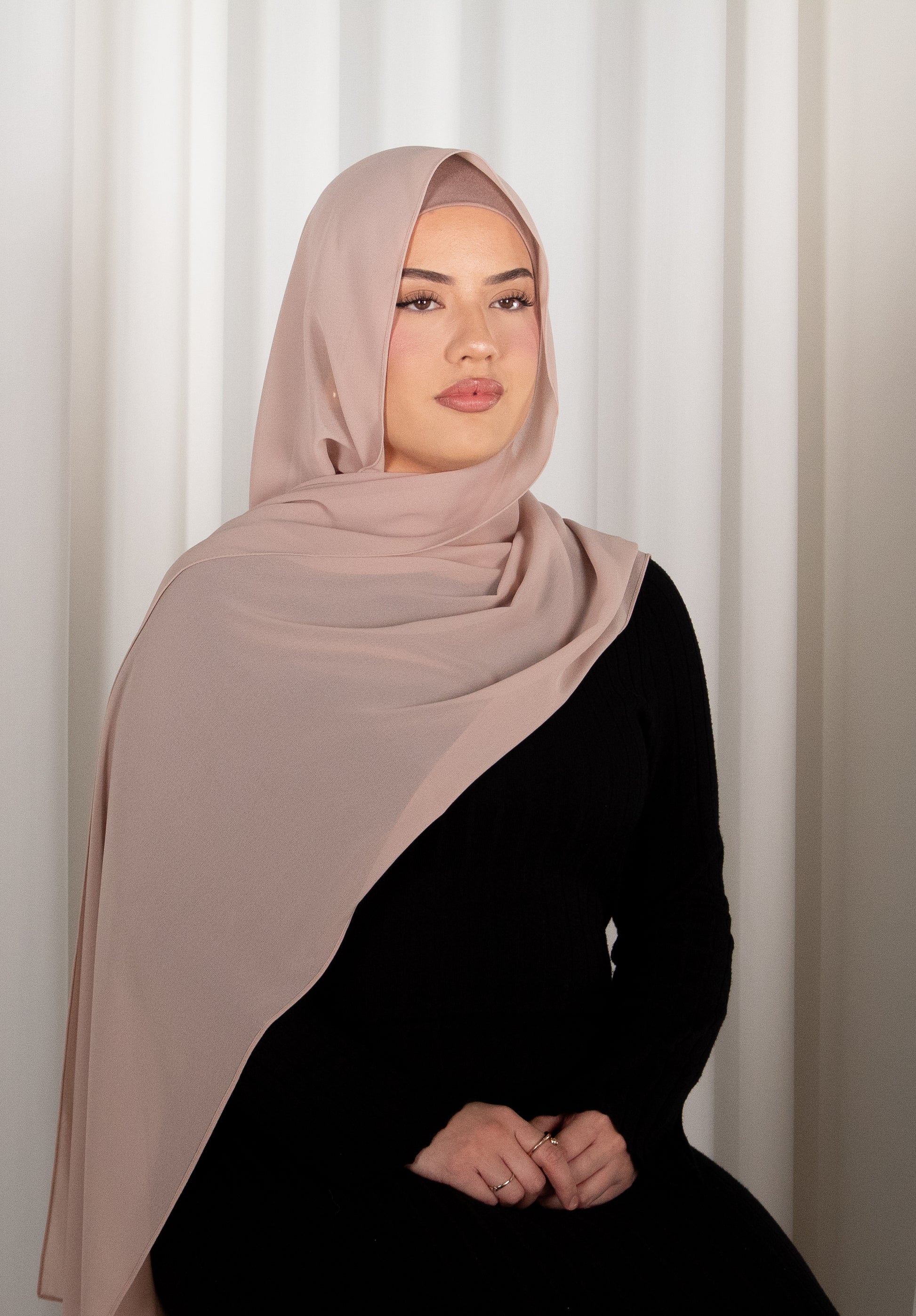 Woman wearing a beige hijab and black outfit against a white curtain background