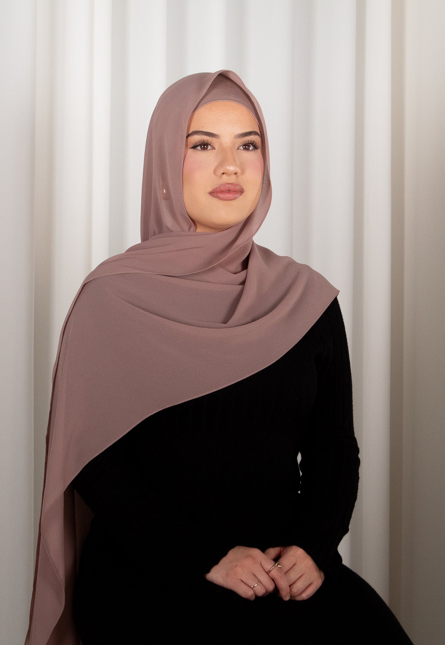 Woman wearing a brown hijab and black outfit against a white curtain background