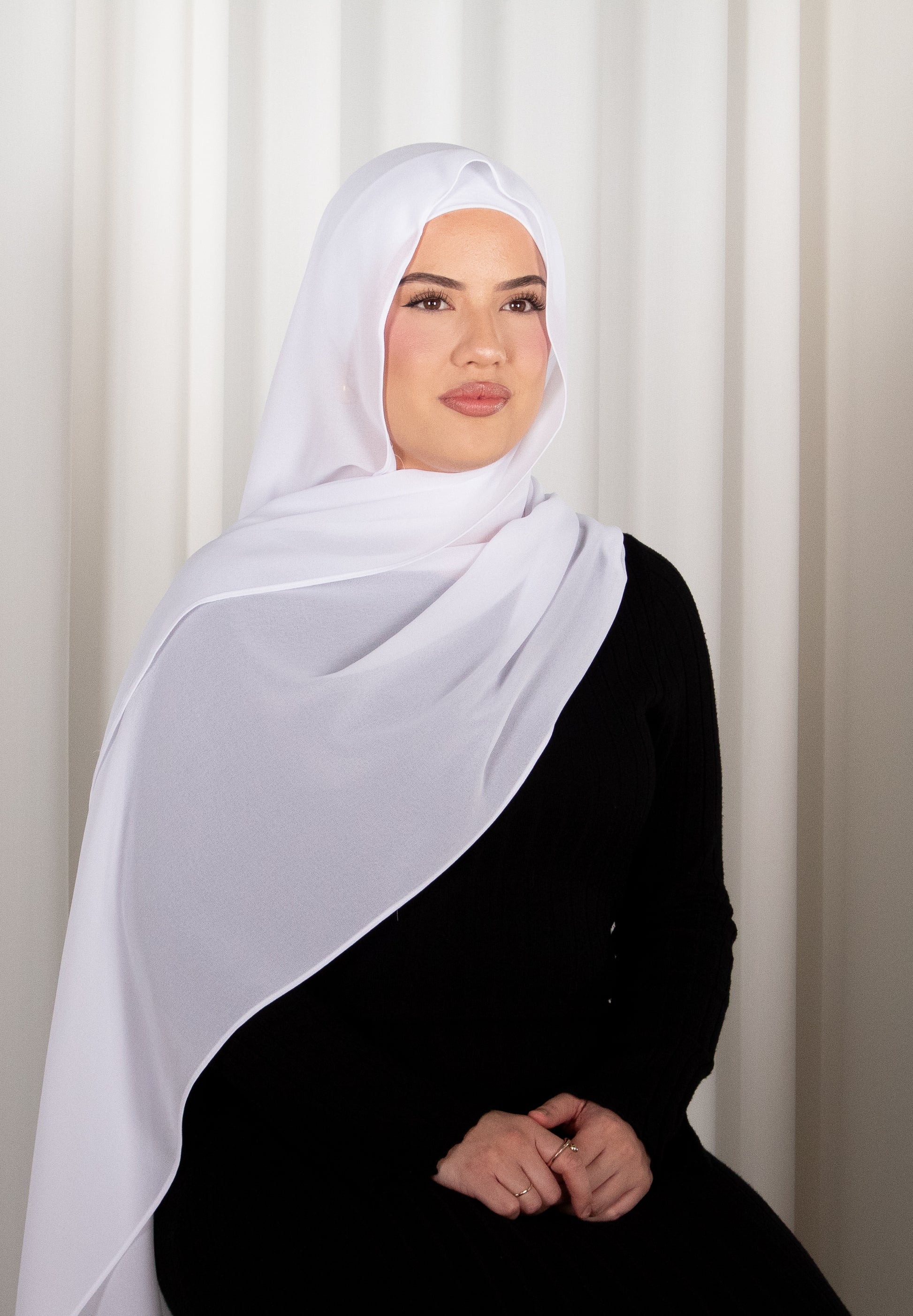 Woman wearing a white hijab and black outfit against a white curtain background