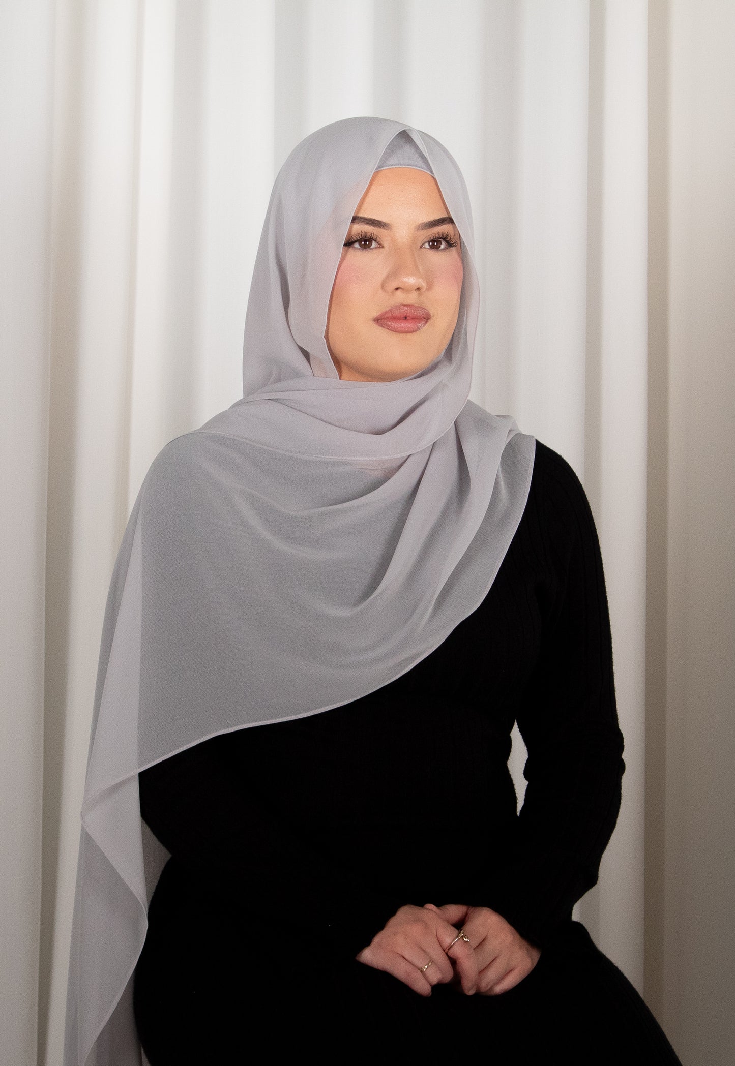 Woman wearing a light gray hijab and black outfit against a white curtain background