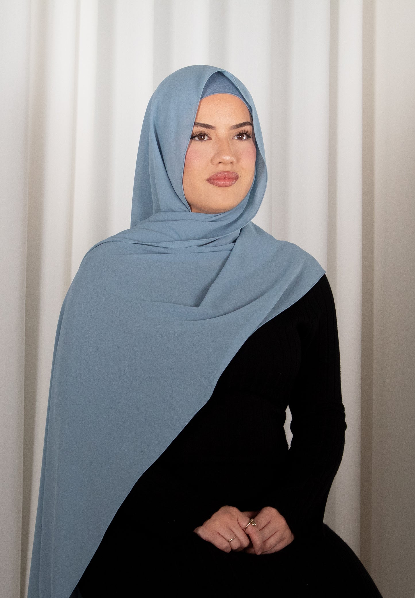 Woman wearing a light blue hijab and black outfit against a white curtain background