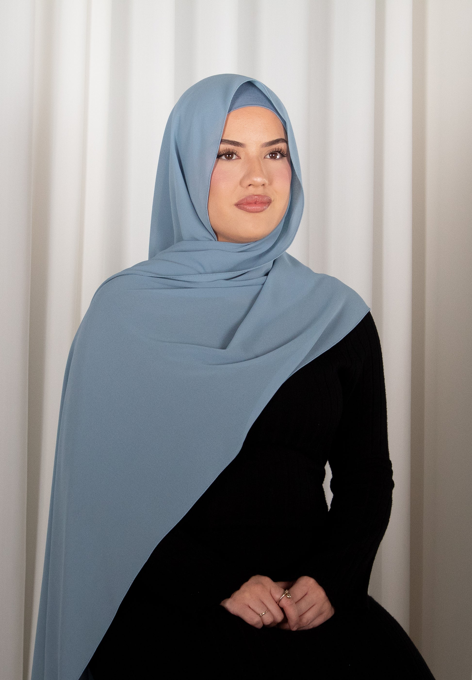 Woman wearing a light blue hijab and black outfit against a white curtain background