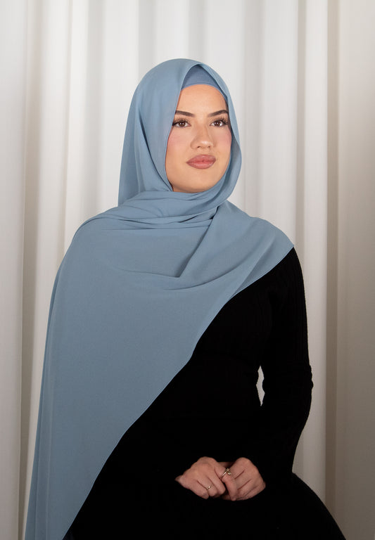 Woman wearing a light blue hijab and black outfit against a white curtain background
