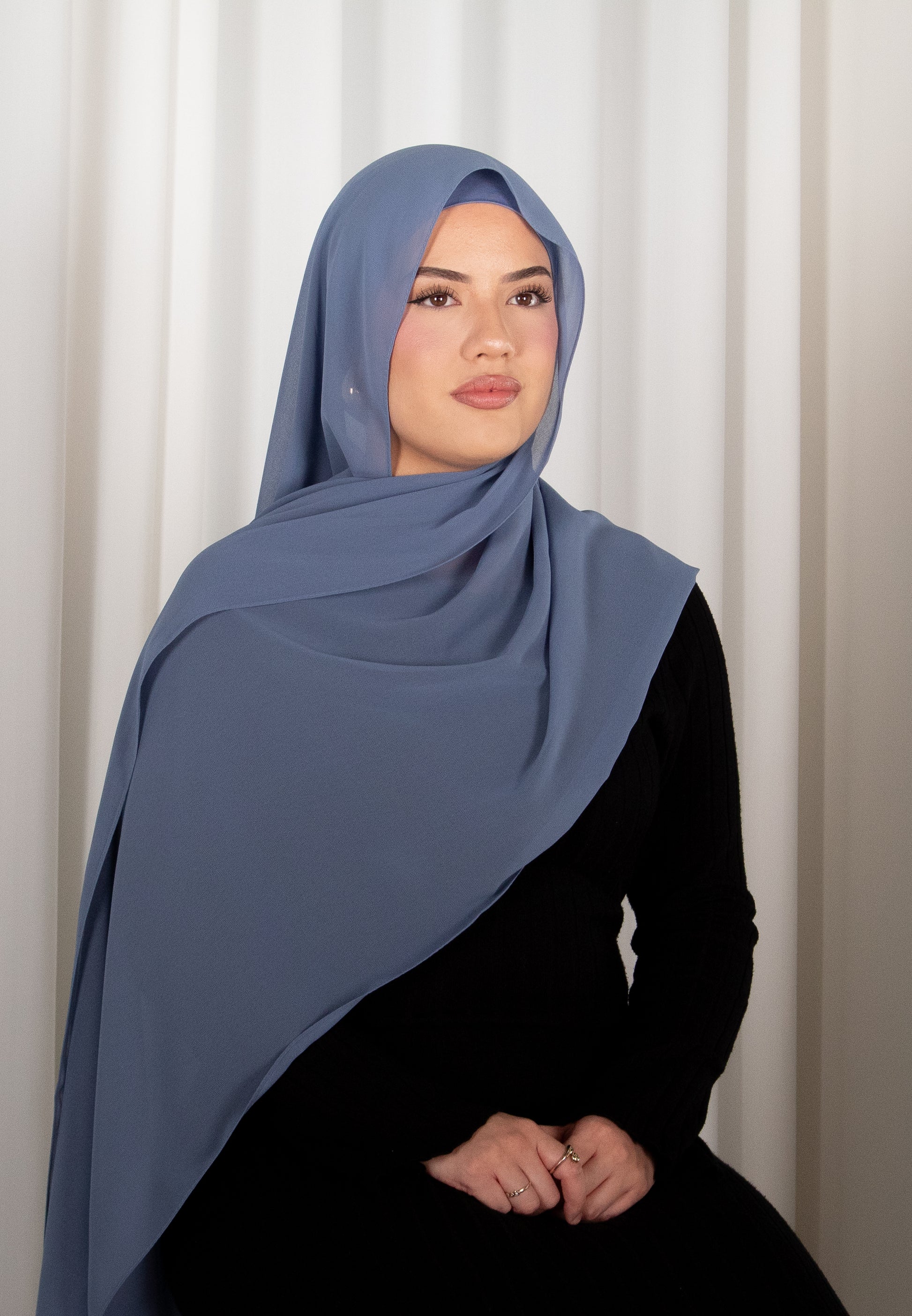 Woman wearing a blue hijab and black outfit against a white curtain background