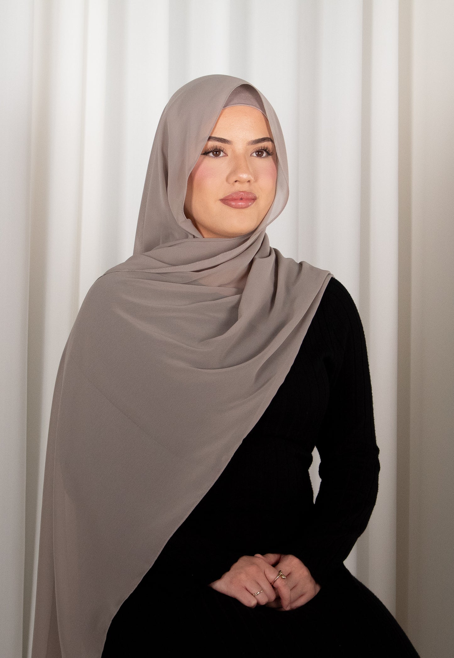 Woman wearing a gray hijab against a white curtain background